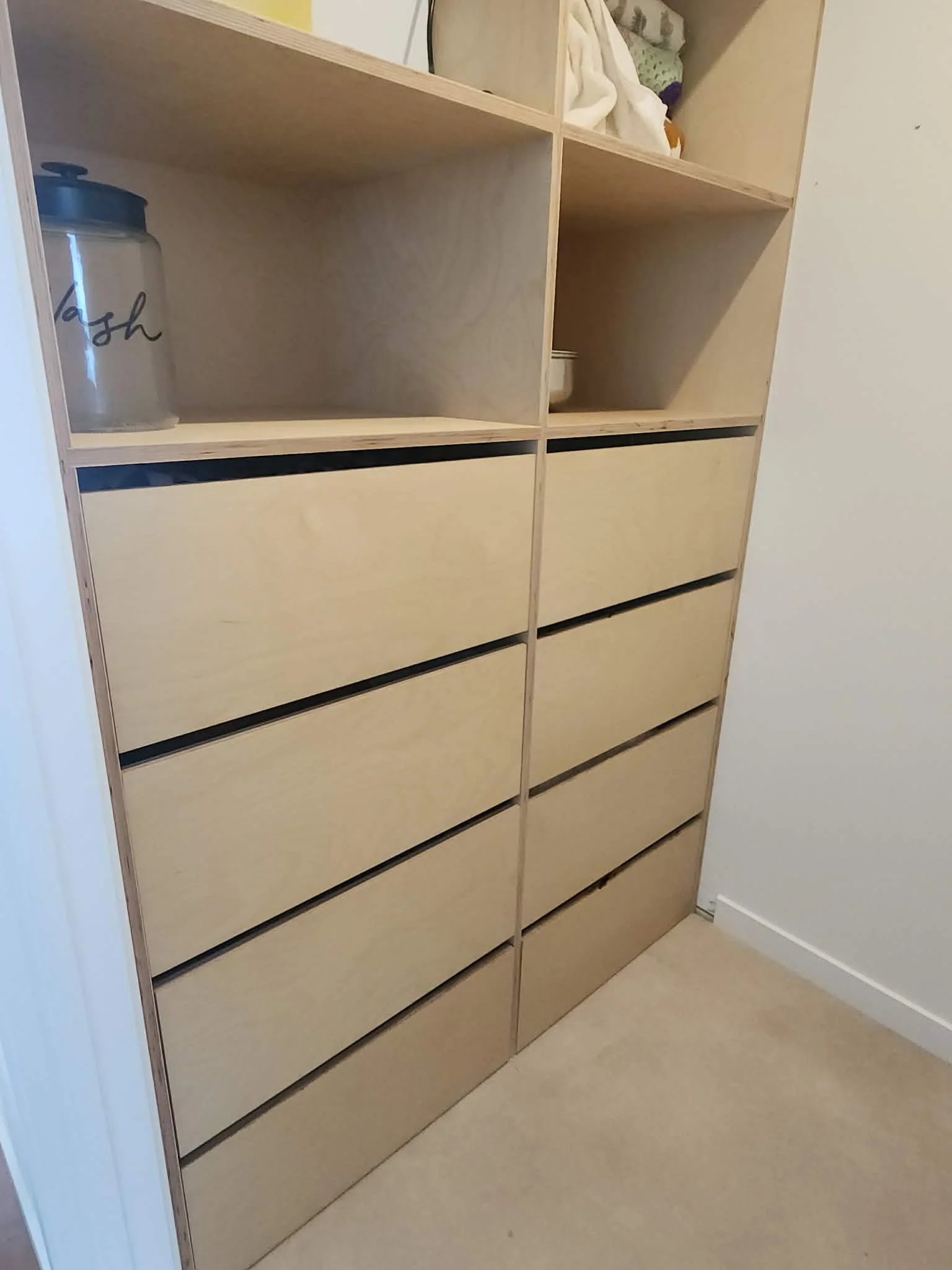 A partially assembled wooden bookshelf with two closed drawers at the bottom and open shelves above, placed in a room with beige carpet and white walls.