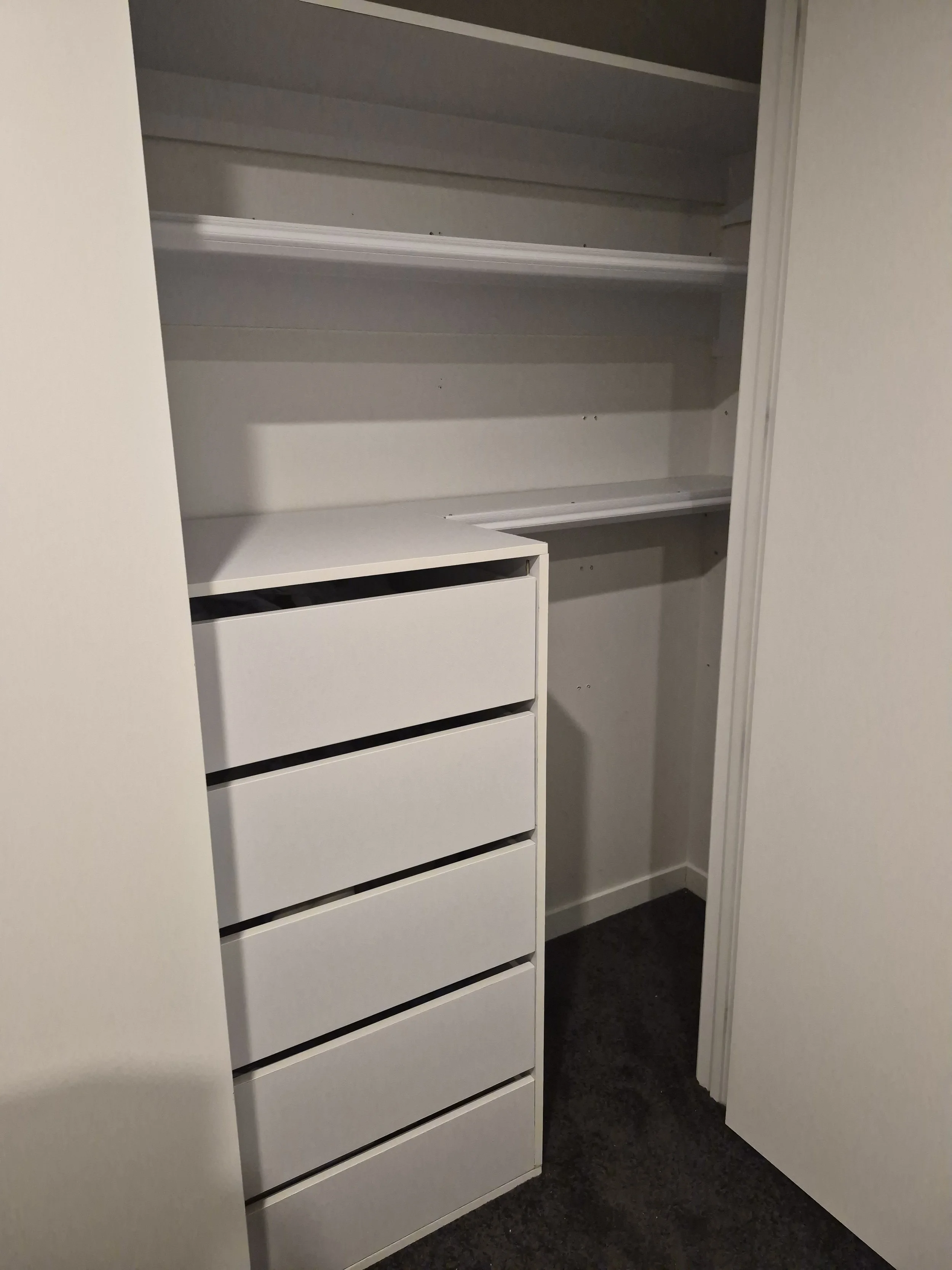 Empty white closet with shelves and drawers, part of a room with a white door and dark carpet.