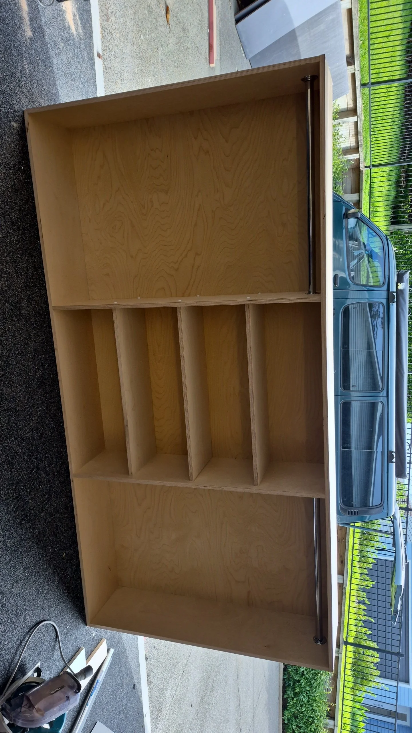 Unfinished wooden bookshelf with five compartments, placed outdoors on a patio, with a car and a black metal fence in the background.