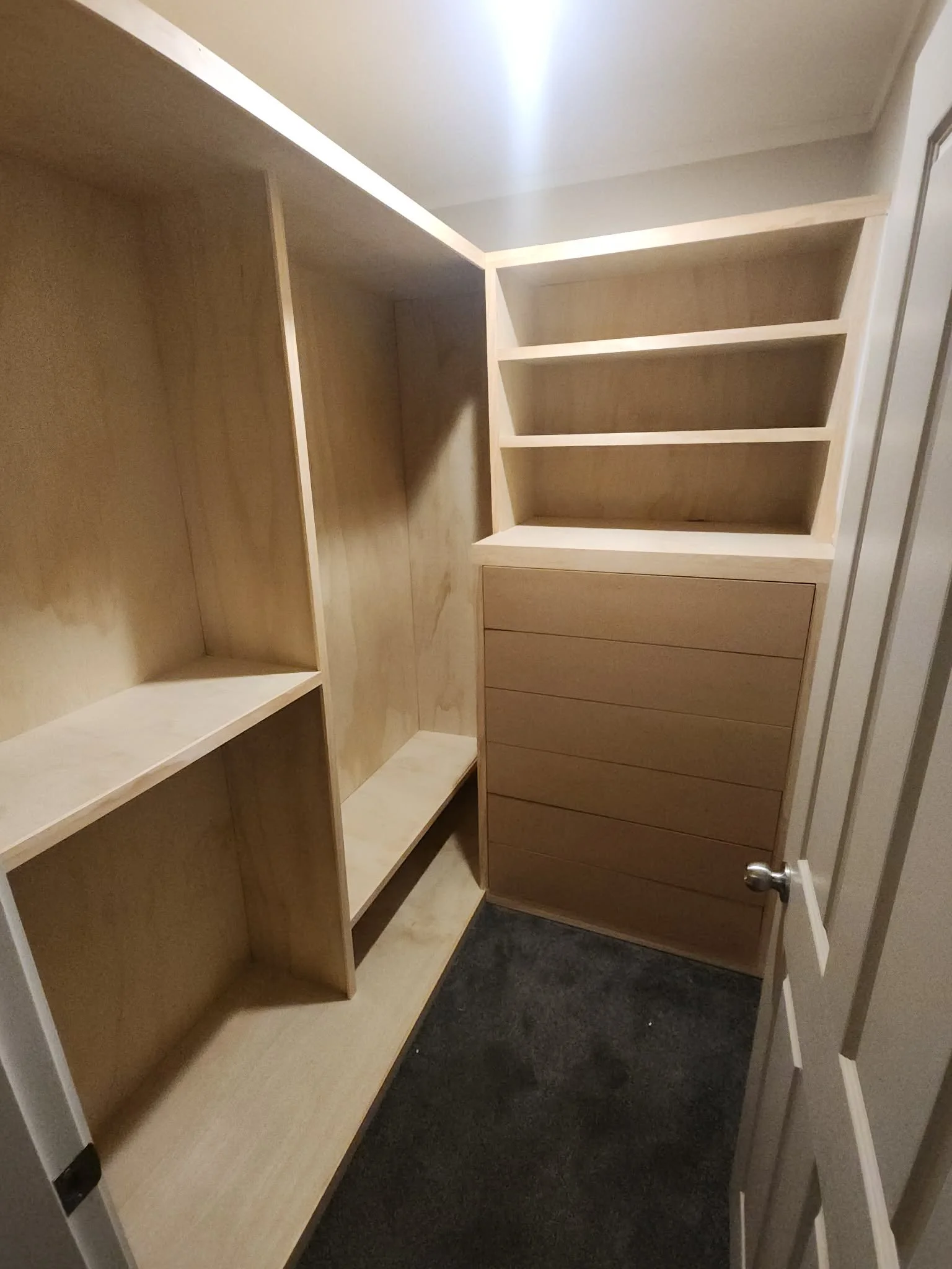 Empty closet with light wood shelves and drawers, beige walls, and dark carpeted flooring.