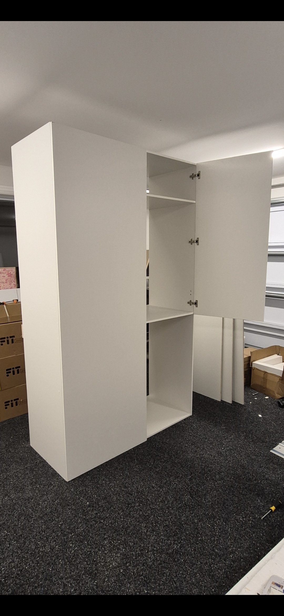 A tall white cabinet with open doors reveals multiple shelves inside, situated in a garage with storage boxes and a rolled-up carpet on a dark carpeted floor.