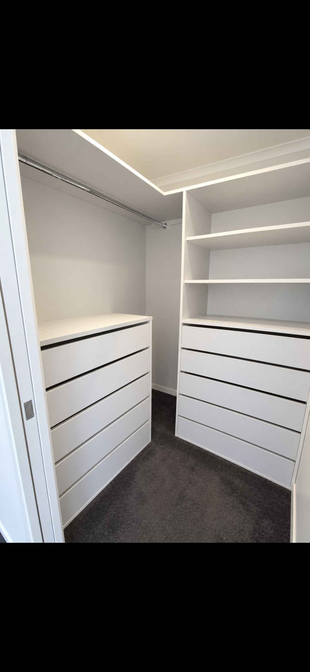 Empty walk-in closet with white built-in shelves and drawers, dark carpeted floor, and a hanging rod for clothes.