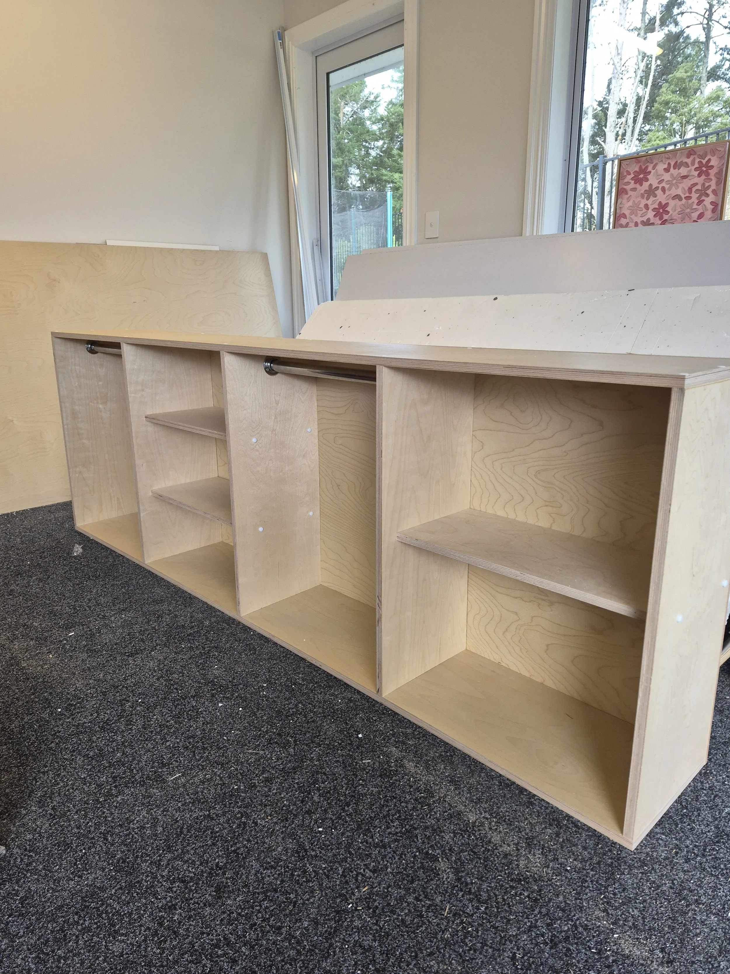 Unfinished wooden bookshelf or storage unit with open compartments placed on a black carpeted floor near a large window.