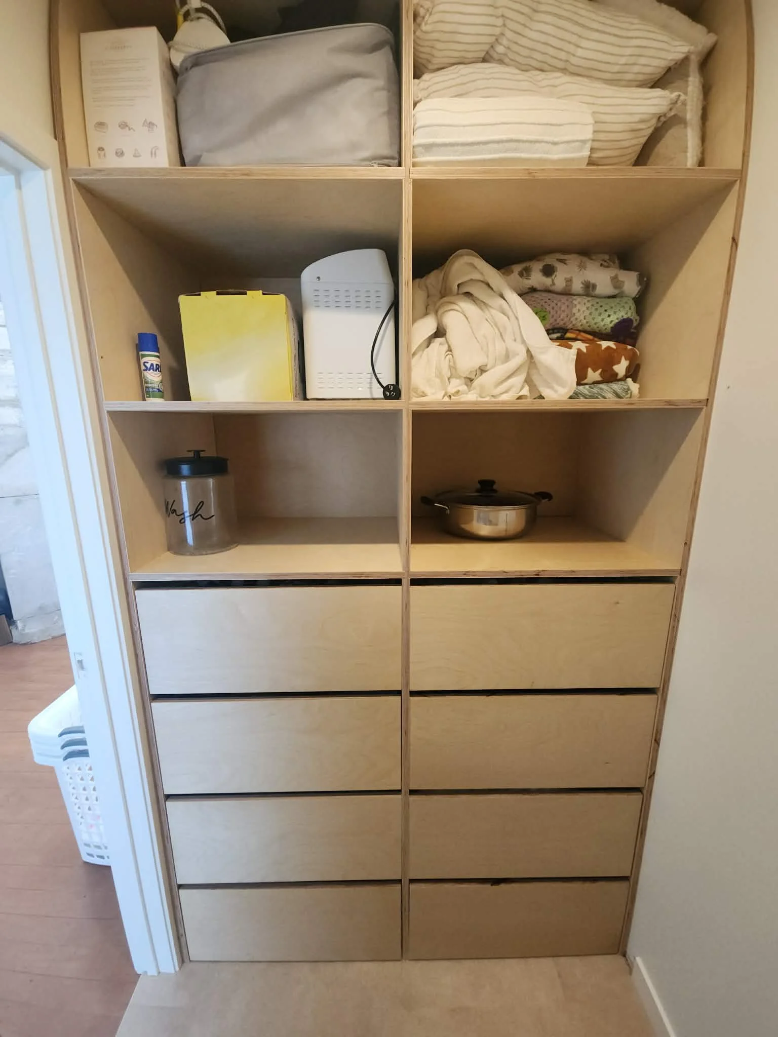 Wooden storage cabinet with multiple compartments holding blankets, a container, a small appliance, and cleaning supplies. Part of the cabinet is empty.