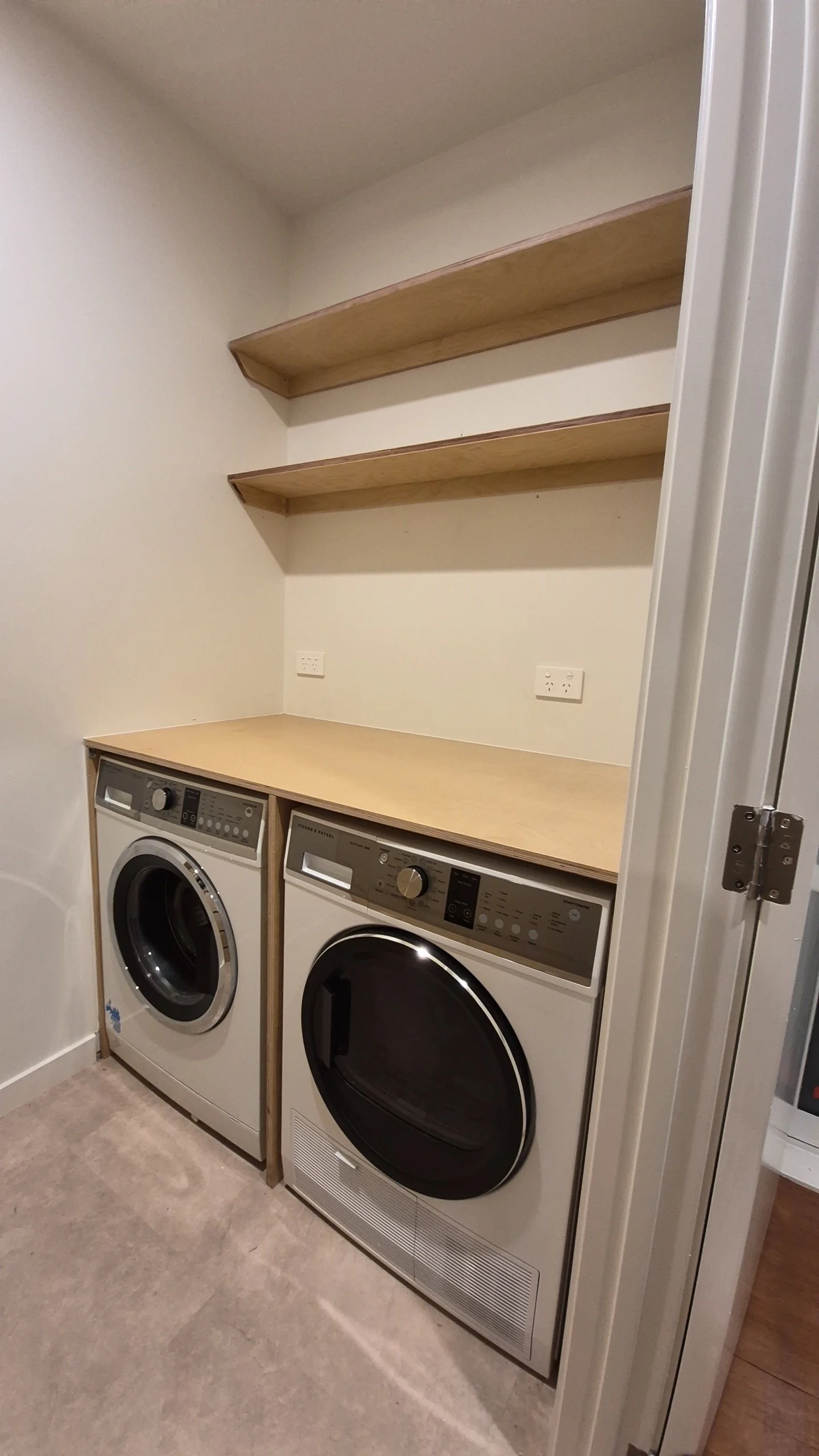 A laundry room with a wooden countertop above two front-load washers and dryers, with two empty wooden shelves mounted on the wall above the countertop.
