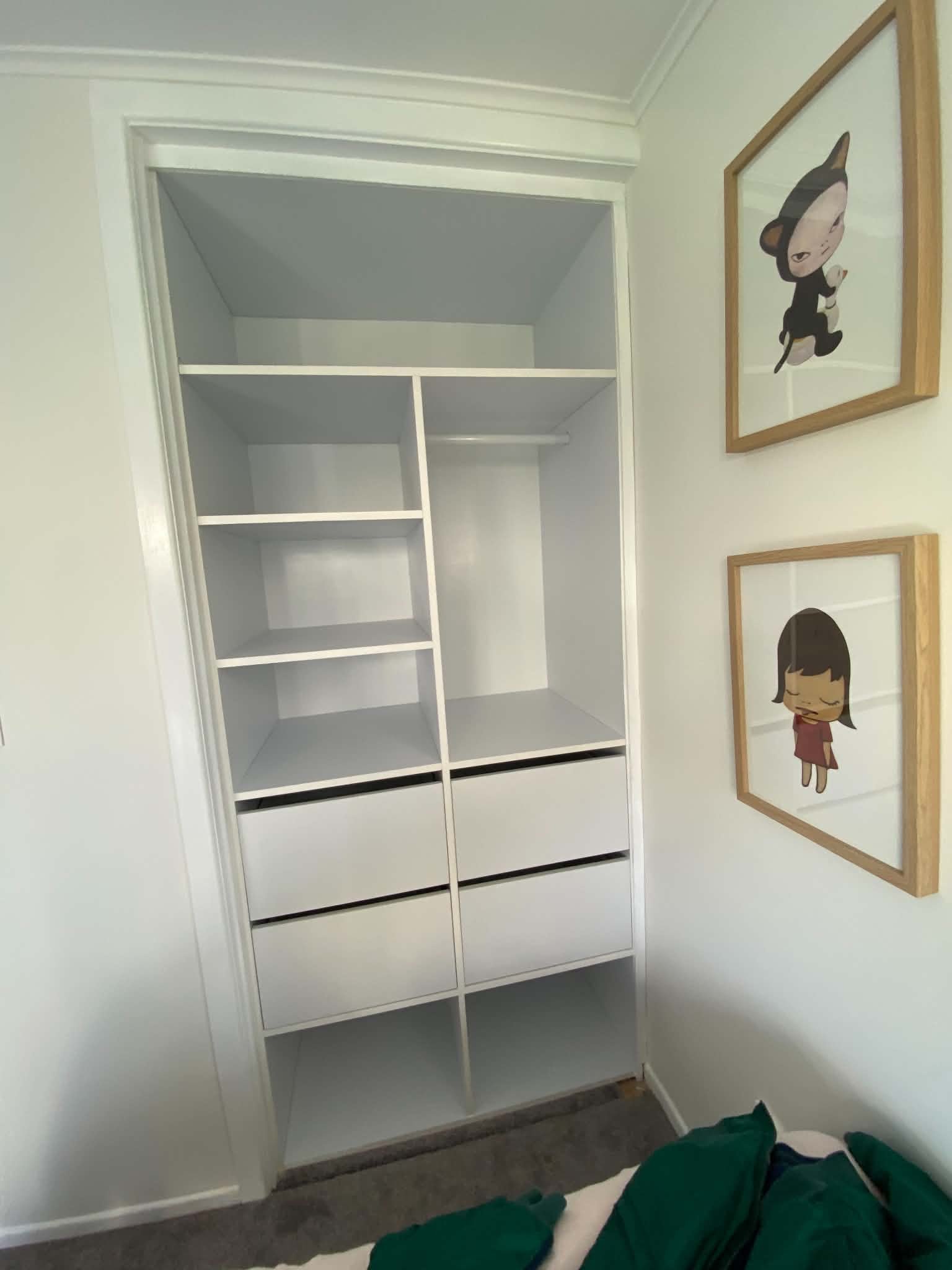 Empty white shelving unit in a corner of a room with two framed cartoon art pictures of children on the adjacent wall, part of a bed with green and black bedding in the foreground, and a carpeted floor.