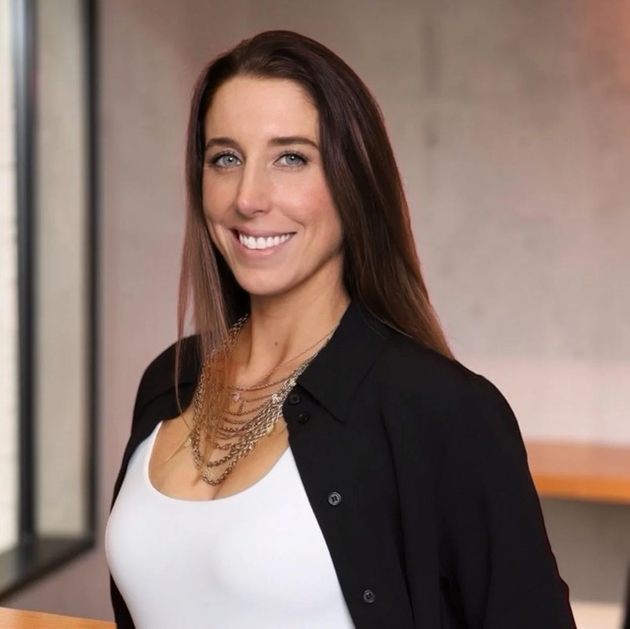 A woman with long brown hair smiling, wearing a black jacket, white top, and layered necklaces, standing indoors with a neutral blurred background.