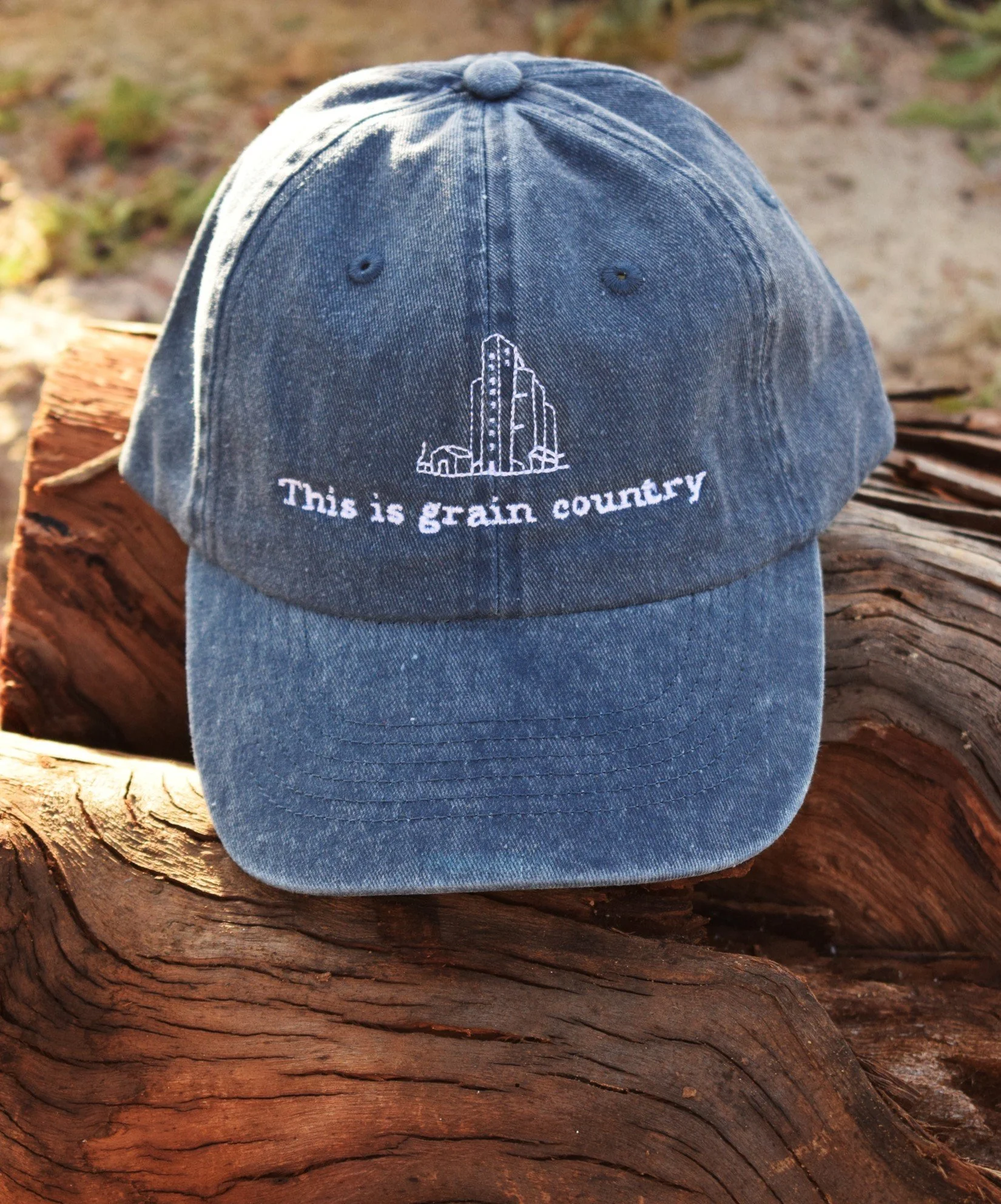 A blue denim baseball cap with white embroidered text and images, resting on a piece of wood.