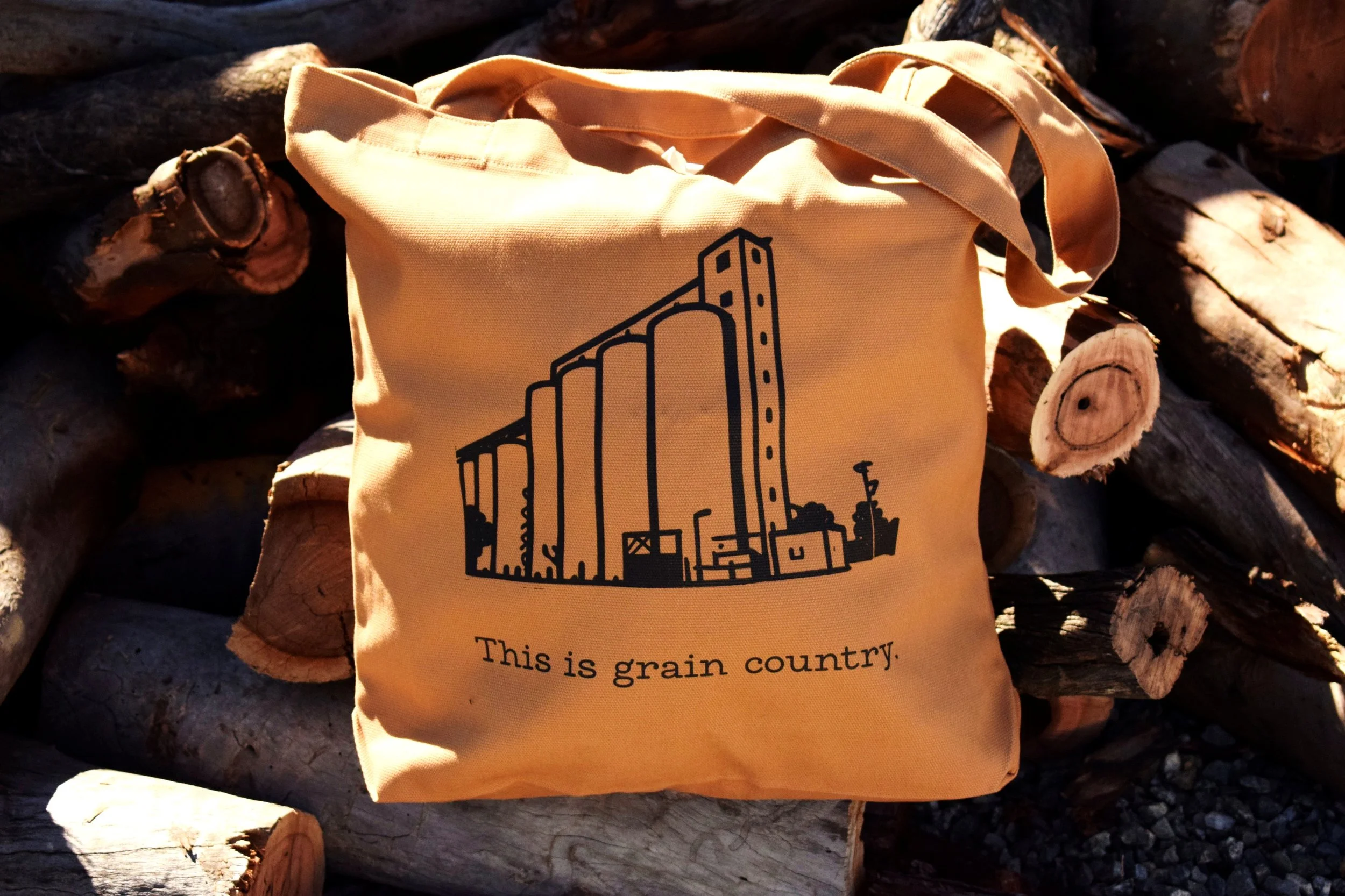 A beige tote bag with a black outline illustration of a grain elevator and the text, "This is grain country," resting atop a pile of chopped wood logs.
