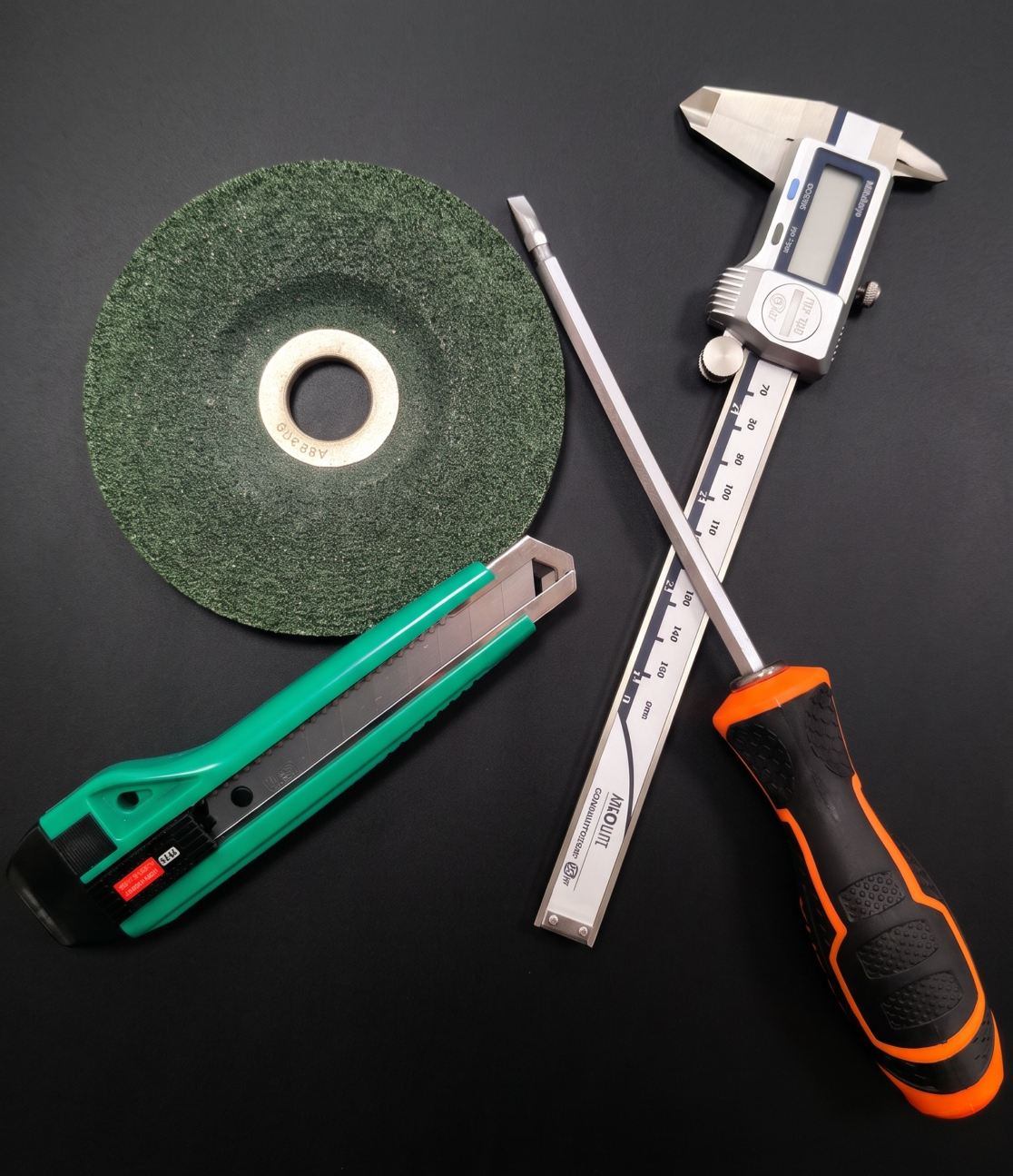 Tools including a grinding wheel, a utility knife, a digital caliper, and a screwdriver with an orange and black handle on a black surface.