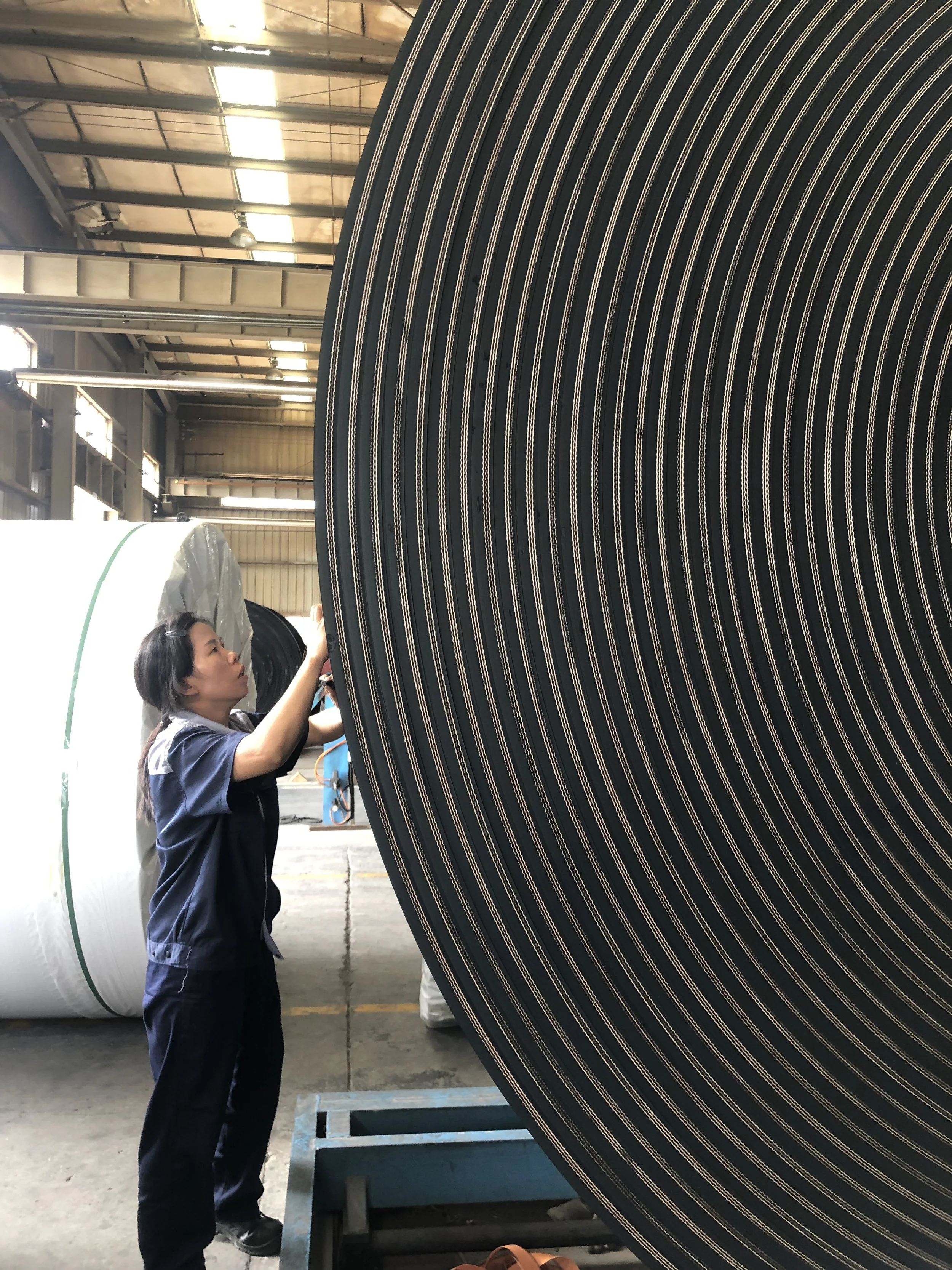A woman in dark clothing standing in an industrial warehouse, inspecting a large conveyor belt with black and beige stripes.