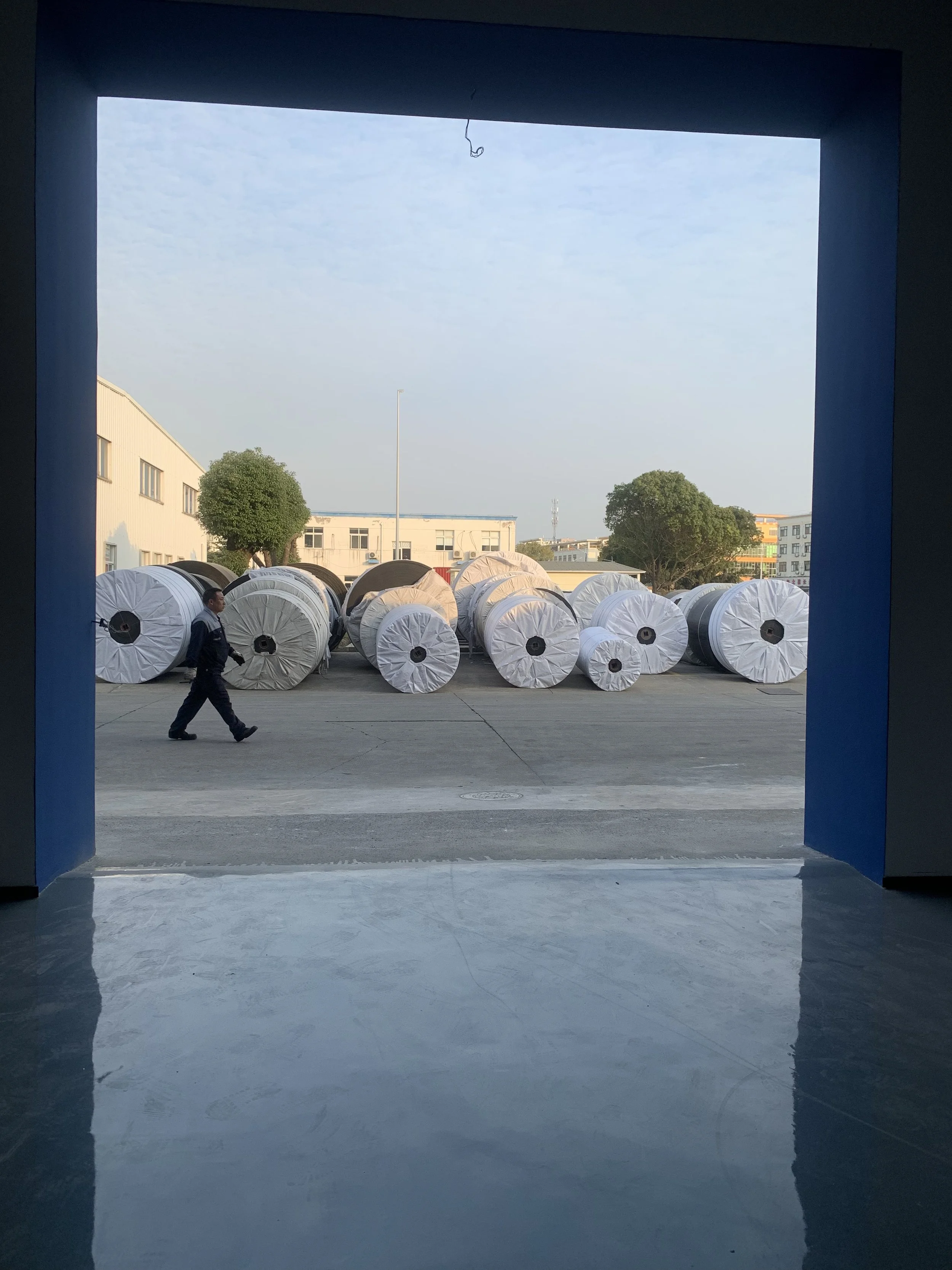 View of large white packaged conveyor belt rolls outdoors through a doorway with bright sky background, a man walking past the rolls, and a few trees and buildings in the distance.