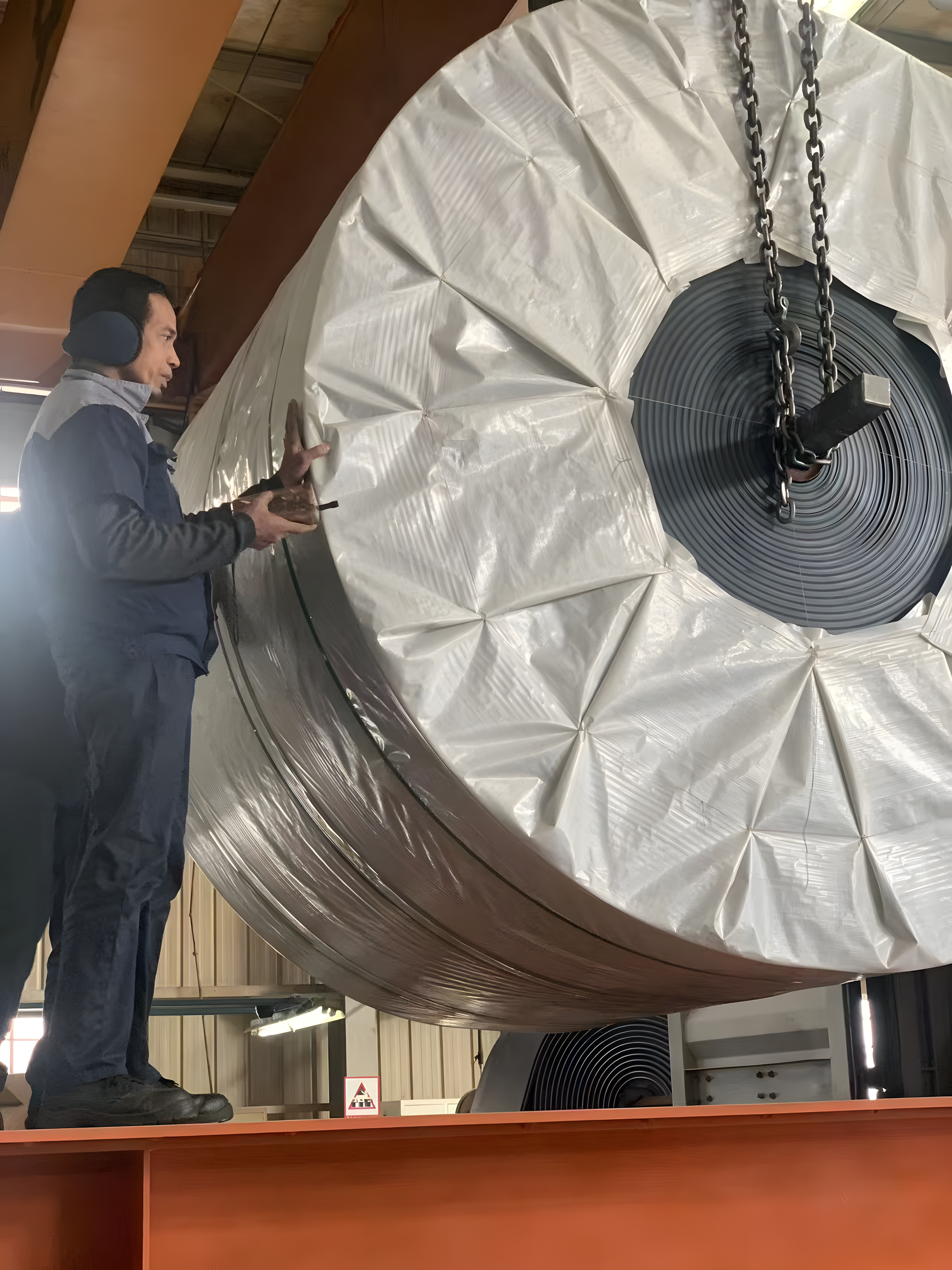 A man wearing earmuffs and work gloves is inspecting a large wrapped industrial conveyor belt with chains attached to it, in an industrial setting.