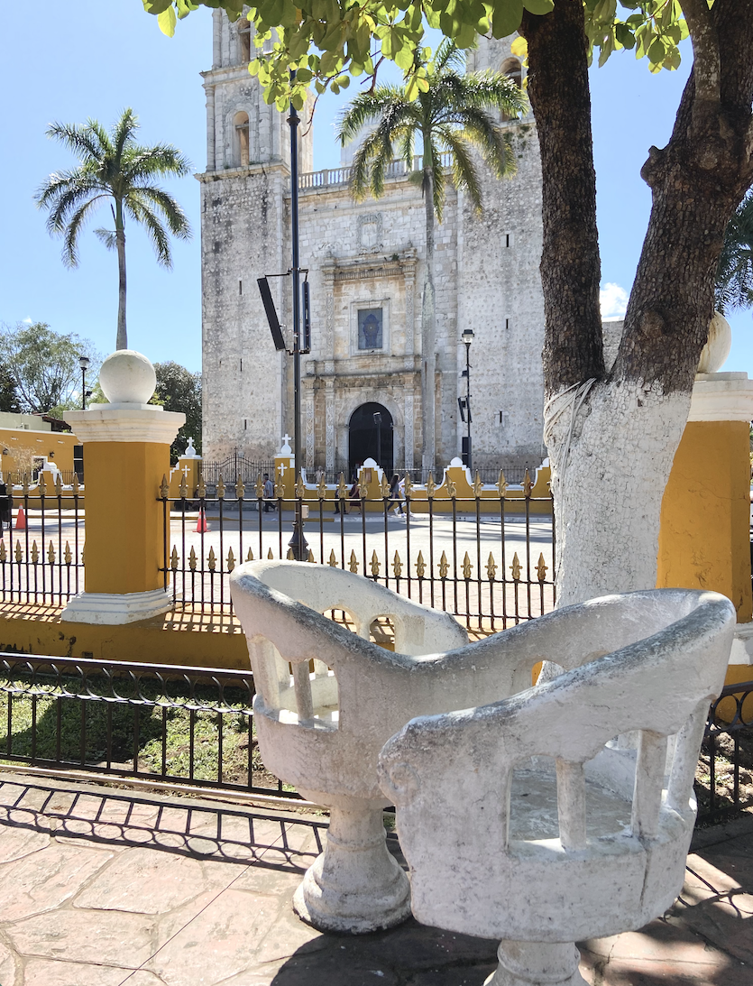 Historic stone church behind a yellow-and-white plaza fence, framed by palm trees and white stone benches under a sunny blue sky, couples together, romantic