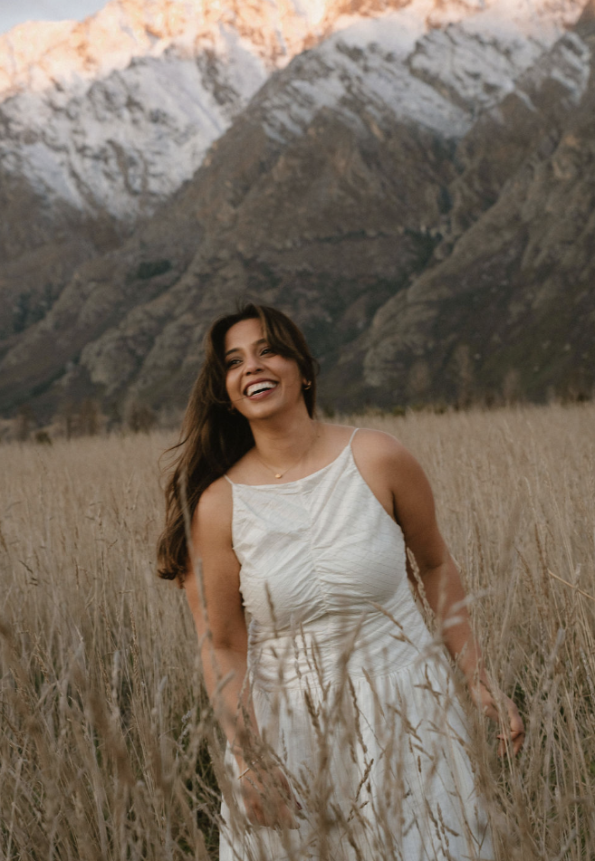 Smiling woman in a white dress standing in a golden grass field with rugged, snow-capped mountains behind her