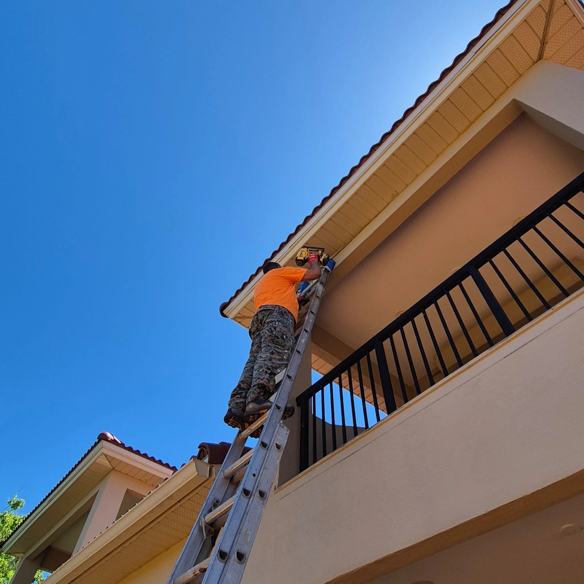 A worker in an orange shirt on a ladder installing or repairing an overhanging roof on a house, with blue sky in the background.