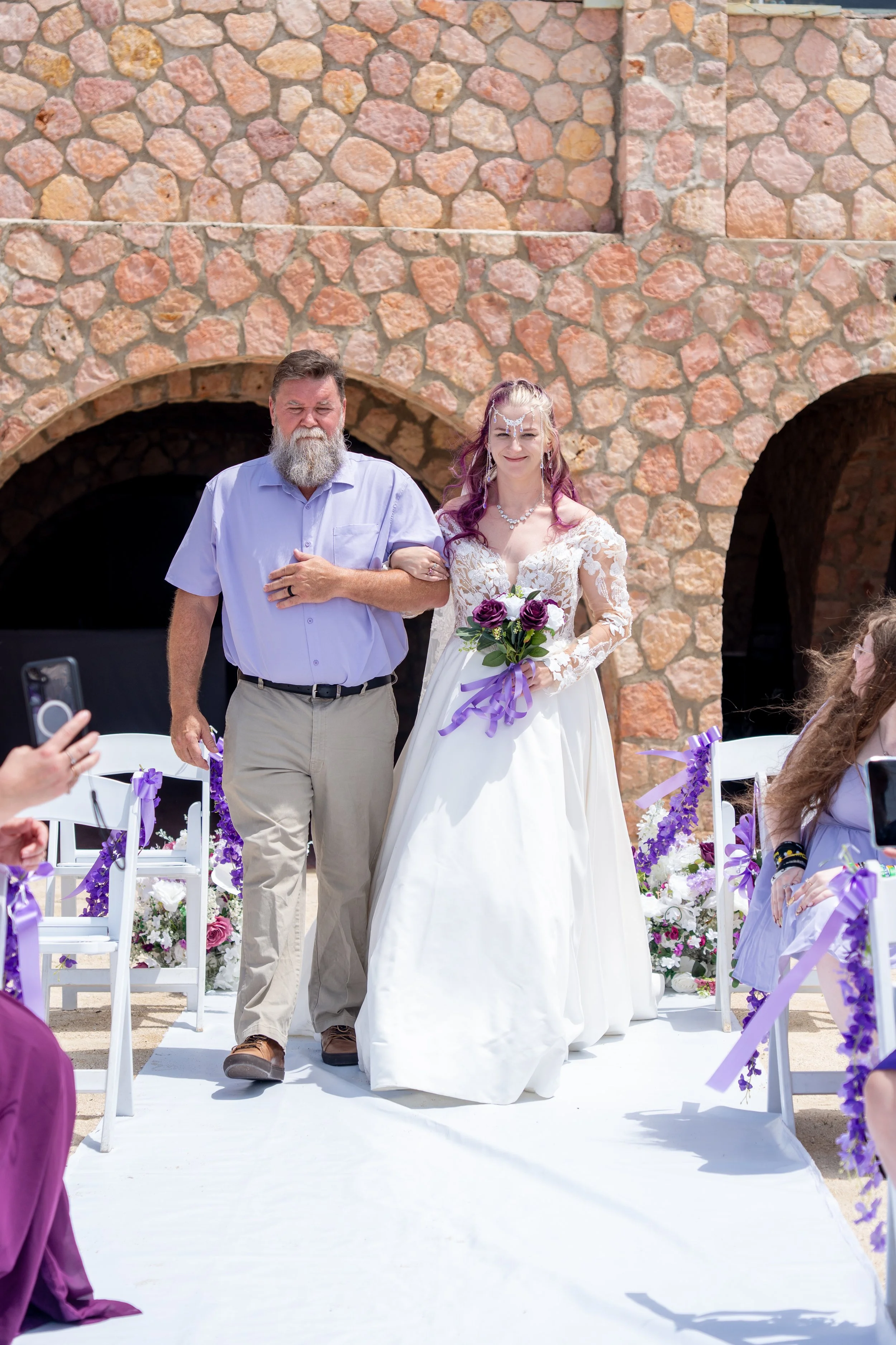 A bride in a white wedding dress holding a bouquet of purple and white flowers walking down the aisle held by a man with a beard wearing a light purple shirt. They are outdoors in front of a stone wall with arches, decorated with purple and white flowers and ribbons.