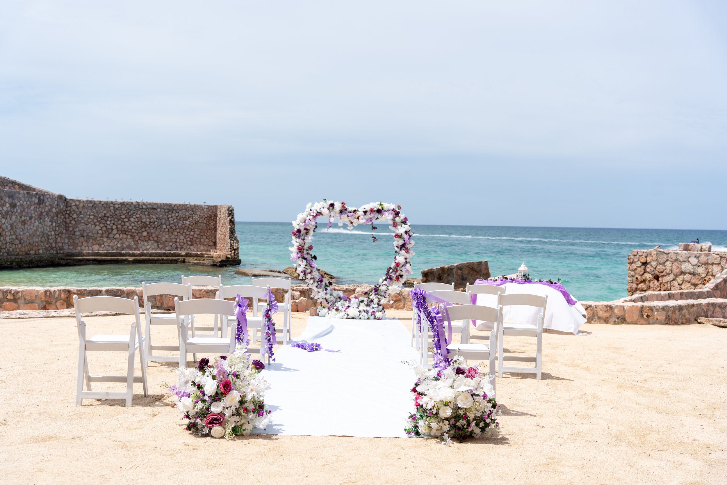 A seaside wedding setup with white chairs decorated with purple ribbons, a floral arch, and flower arrangements, overlooking the ocean with a breakwater wall on the left and a cloudy sky.