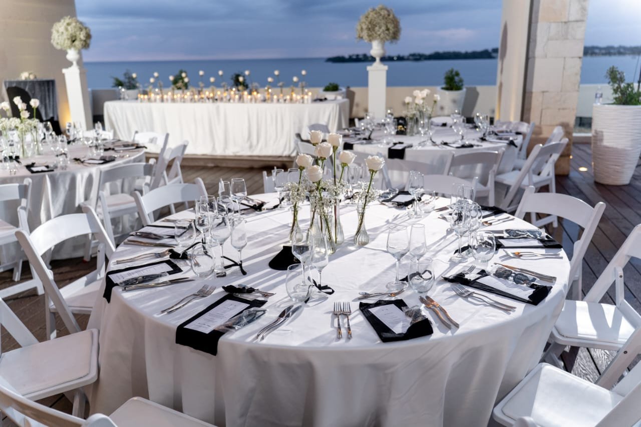 Elegant outdoor wedding reception setup with round tables covered in white tablecloths, decorated with white roses and greenery centerpieces, surrounded by white chairs, overlooking a body of water during sunset.