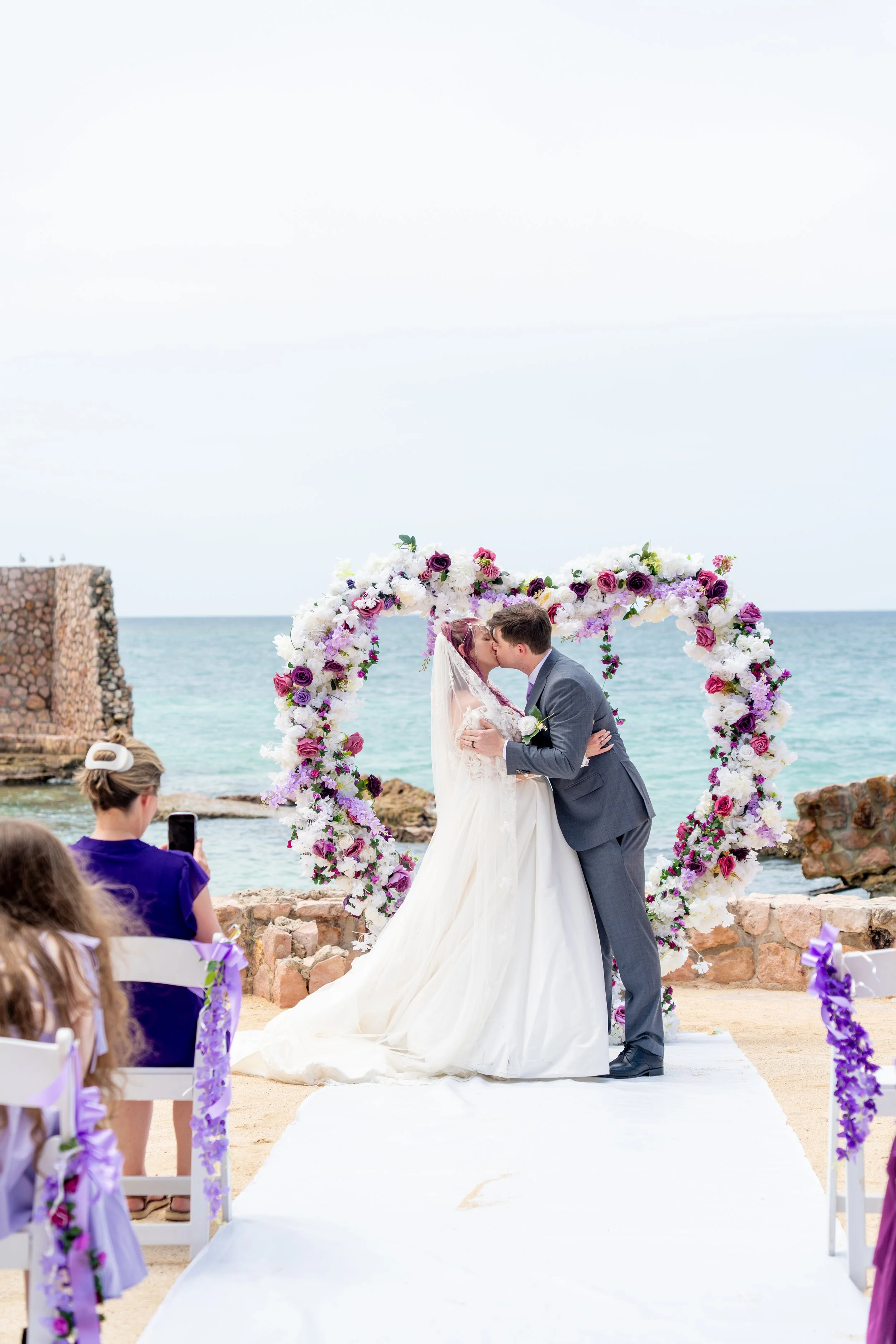 A bride and groom kiss underneath a floral wedding arch by the ocean, with guests seated nearby.