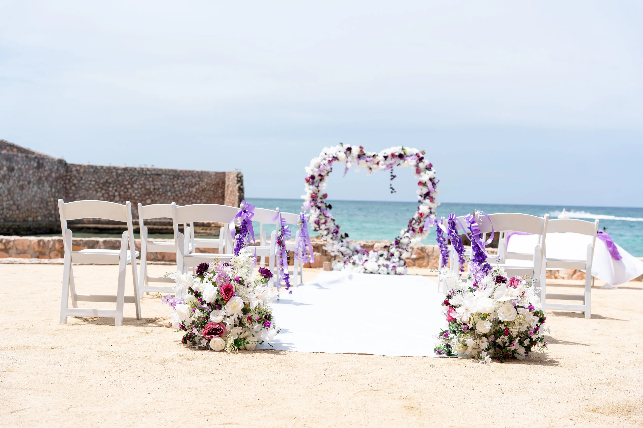 Beachside wedding setup with a floral arch, white chairs, and flower arrangements on a sandy beach, overlooking the ocean with a cloudy sky.