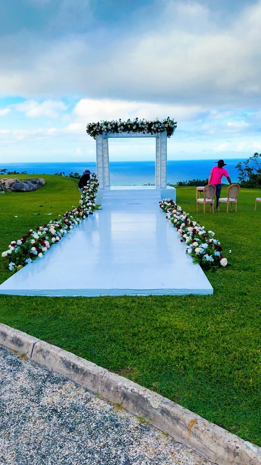 Outdoor wedding ceremony setup with an aisle decorated with flowers leading to a floral arch, overlooking the ocean.