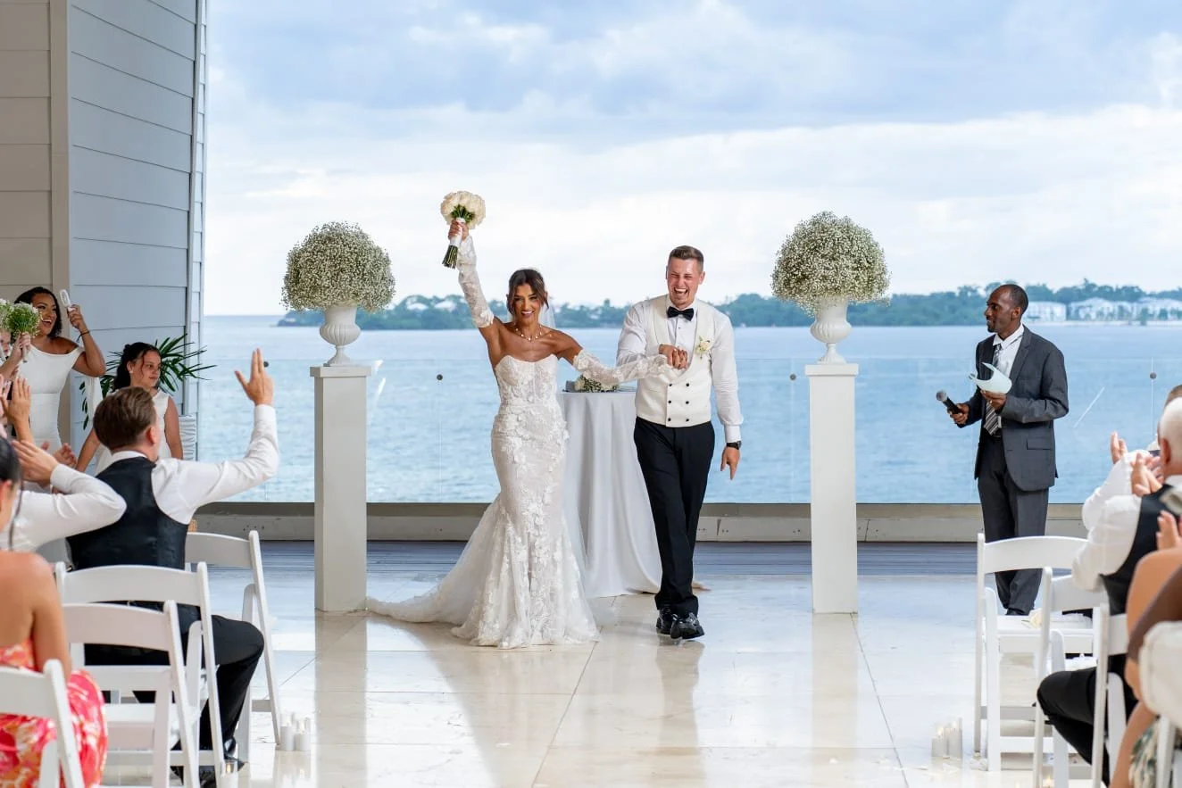 Bride and groom walking down the aisle at their wedding ceremony with guests applauding and celebrating, set against a waterfront view.
