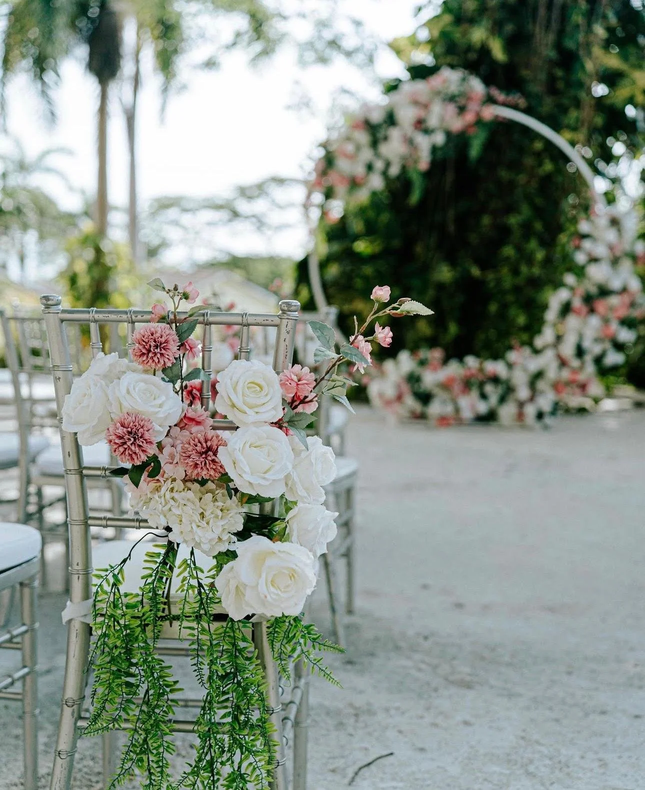 Flower arrangement with white roses, pink dahlias, and greenery hanging on a silver chair at an outdoor event, with a blurred floral arch in the background.