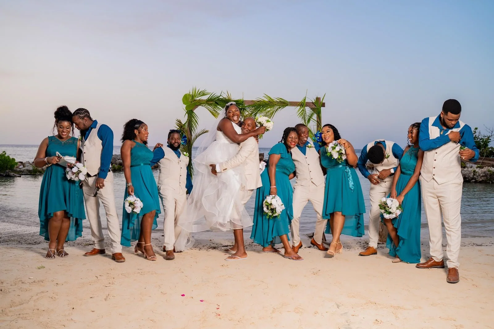 A wedding party on a beach with the ocean in the background. The bride, in a white dress, is being lifted by the groom, and they are surrounded by bridesmaids and groomsmen in teal and cream outfits. The group is happy and celebrating.