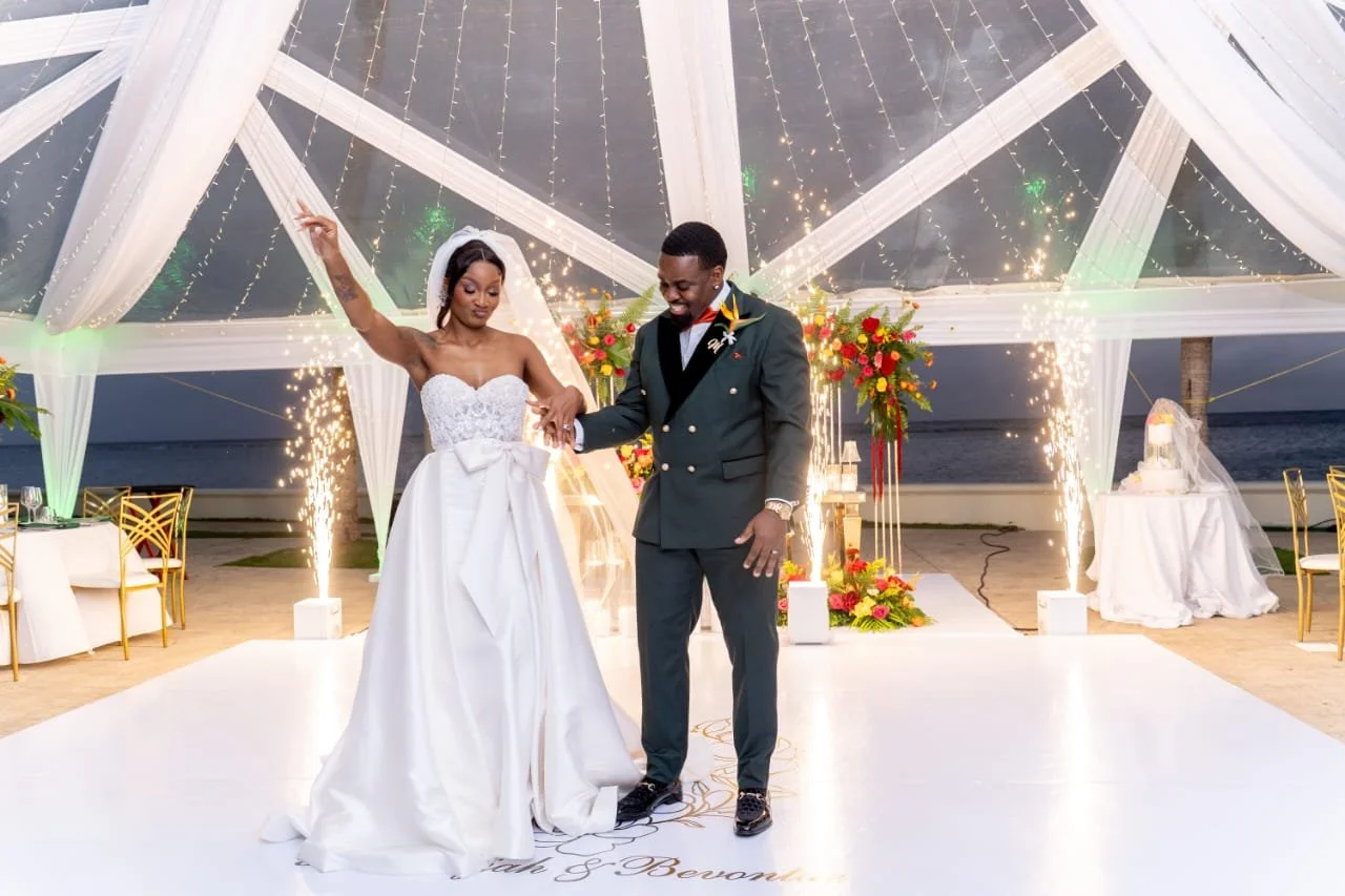A bride and groom dancing under a decorated outdoor wedding canopy at sunset, with sparklers and fireworks.