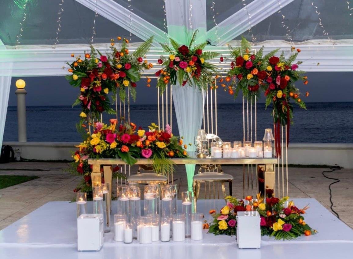 A decorated table with vibrant flowers, candles, and glassware on a white tablecloth, set outdoors near a body of water with a tent and sheer drapes overhead.