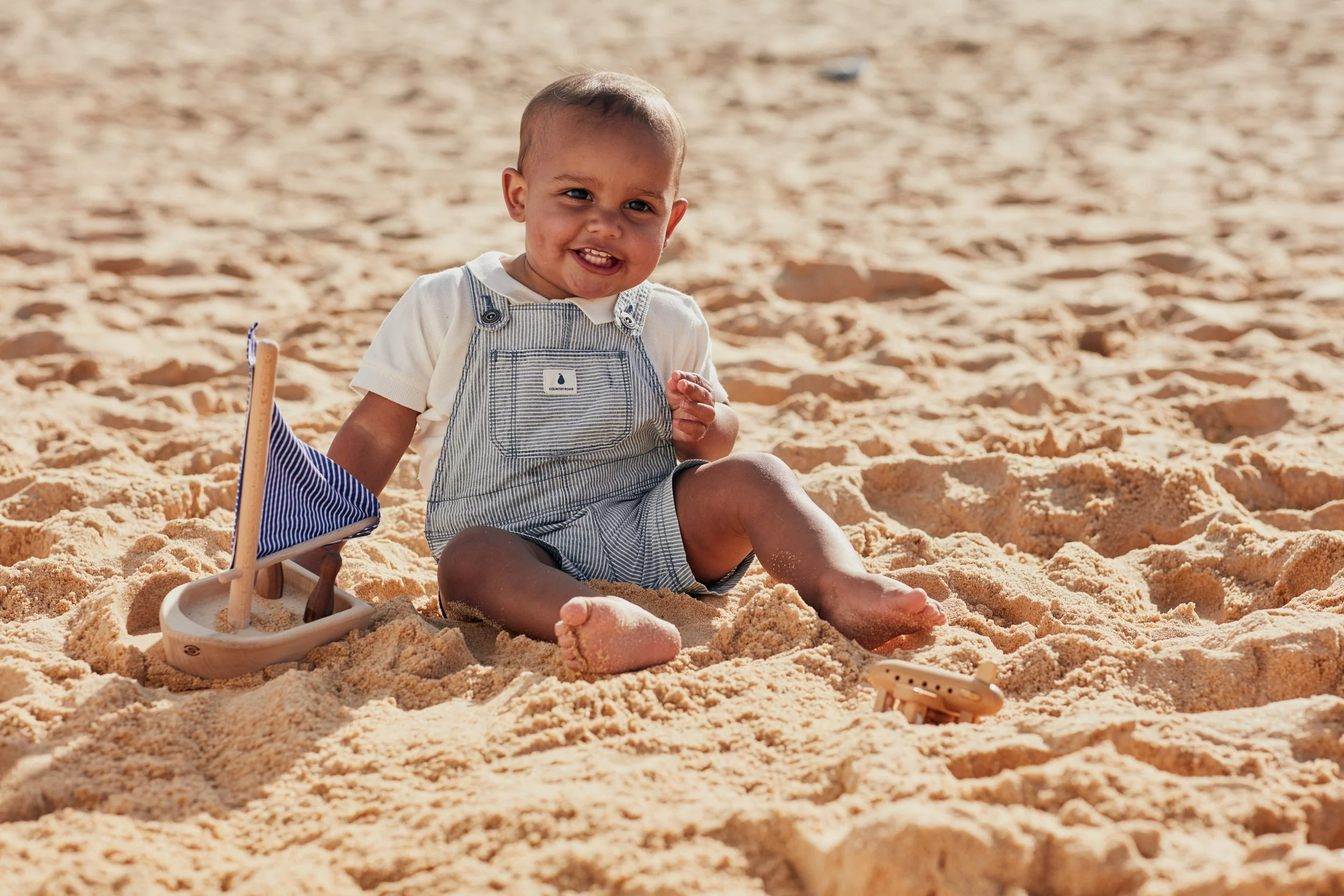 Smiling baby in striped overalls sitting in a sandbox with beach toys