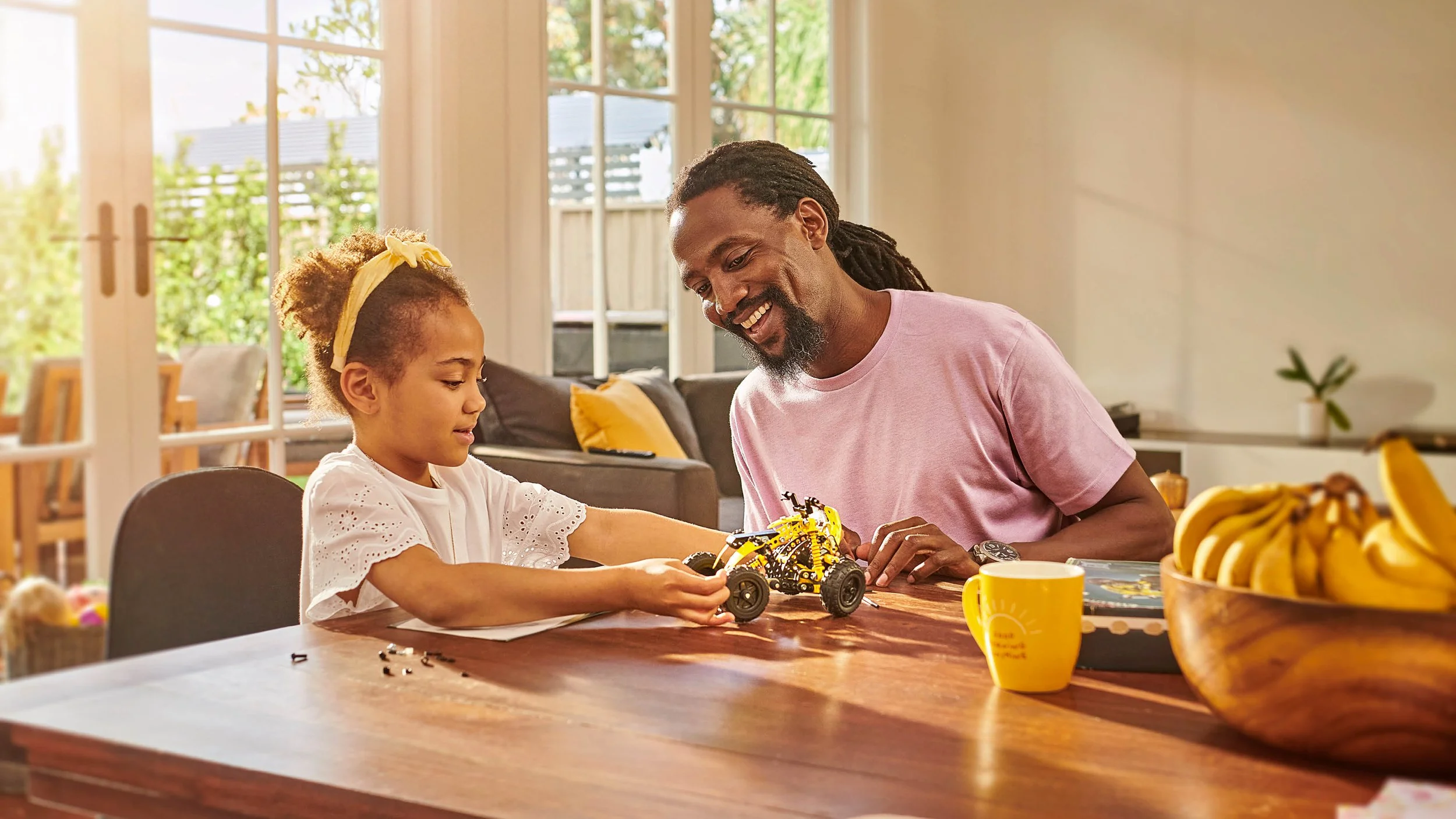 Father and daughter smiling while building a toy car at a sunlit dining table
