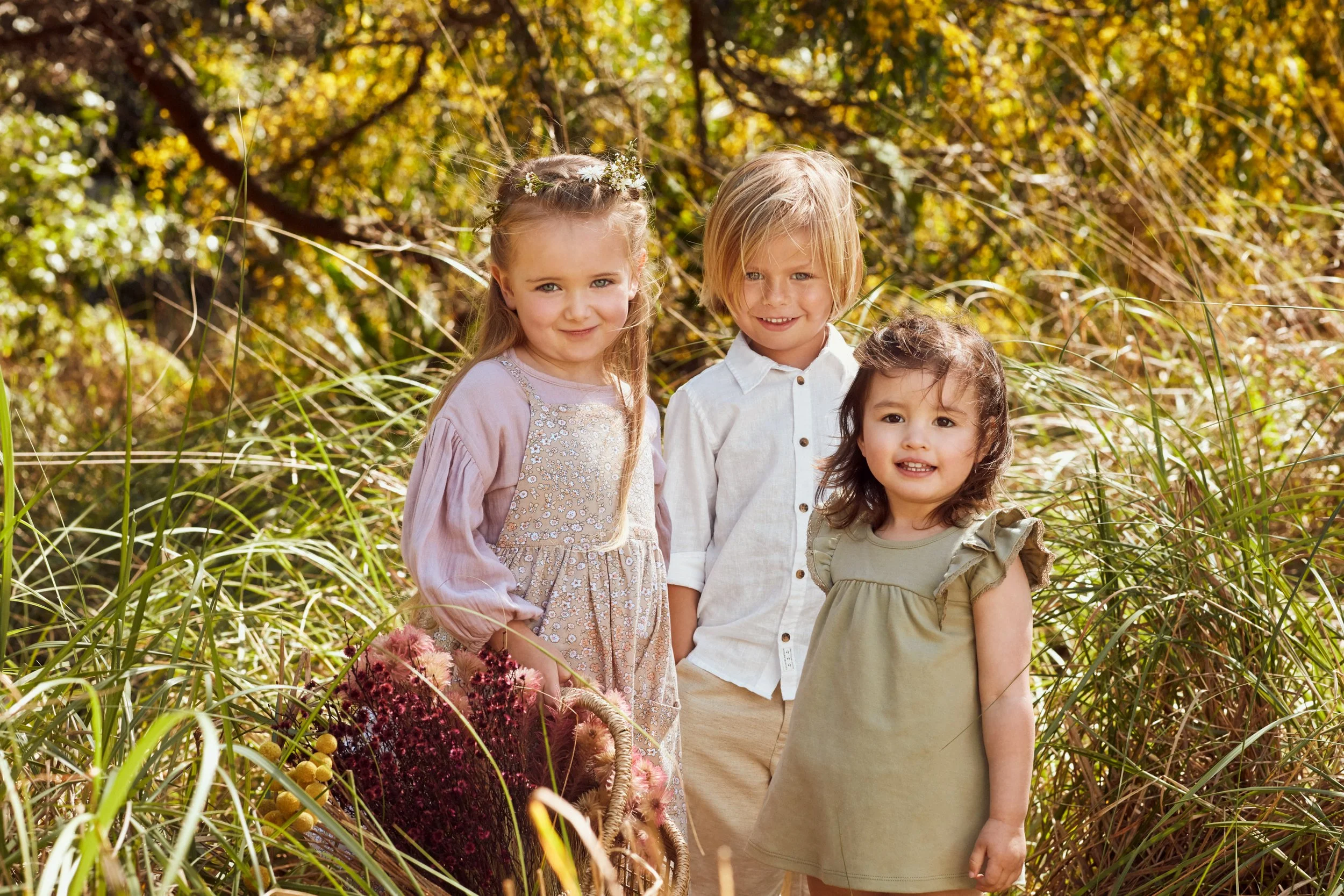 Three children smiling outdoors in a sunlit meadow, surrounded by tall grass and autumn trees.