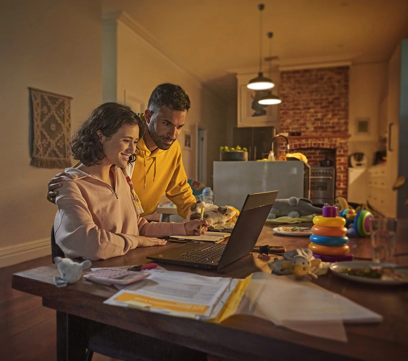 Parents helping a child with homework on a laptop at a kitchen table surrounded by toys and papers.