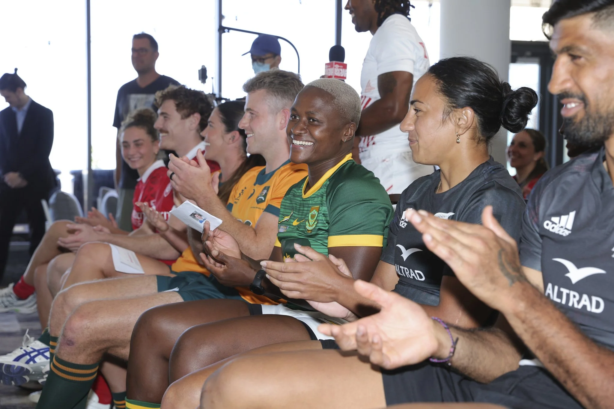 Diverse group of athletes applauding during an indoor event, including a smiling woman in a green and yellow sports jersey.