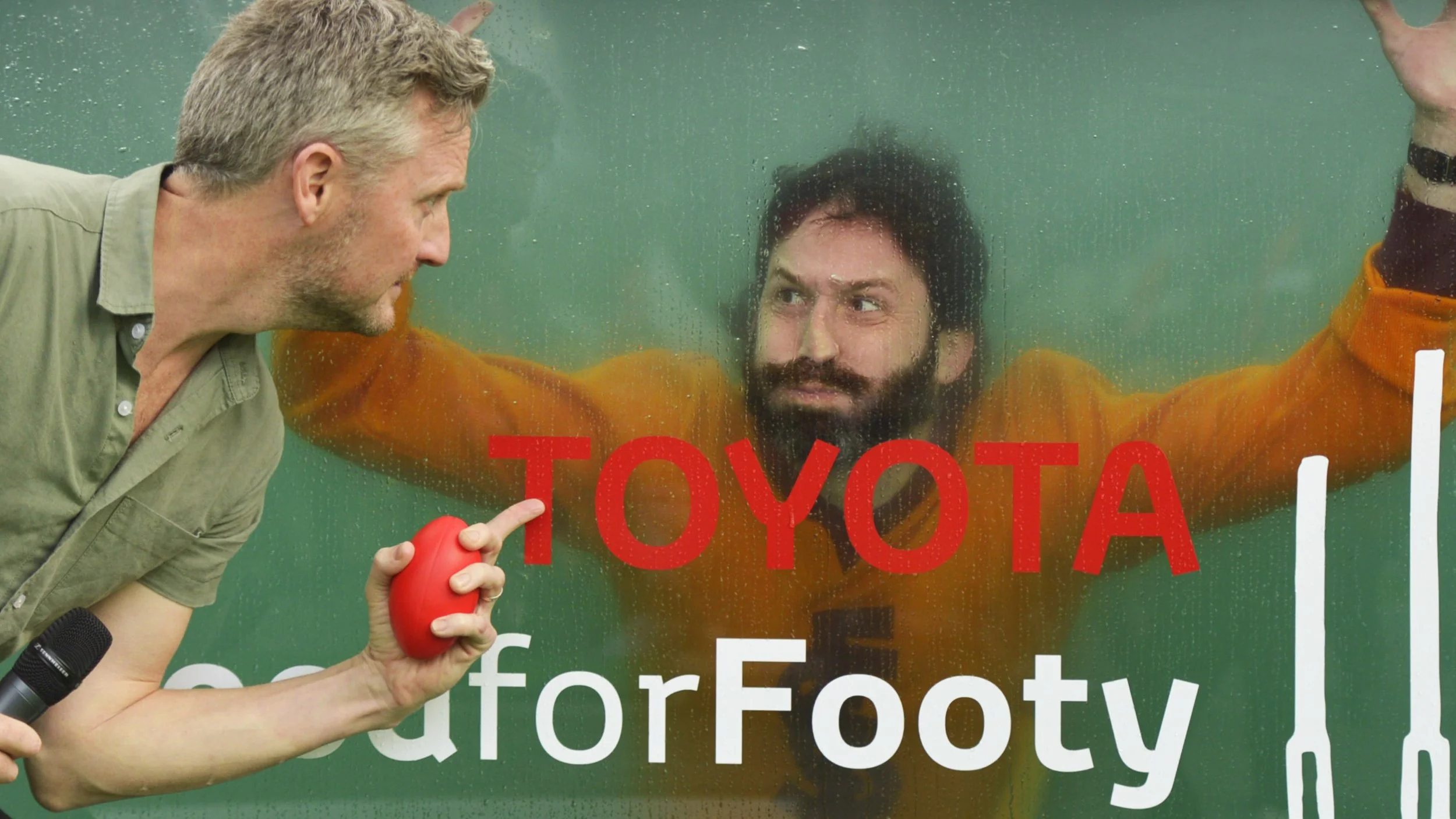 Man holding a red ball interviews a bearded man behind wet glass in an orange prison jumpsuit, with “TOYOTA” and “for Forty” text visible.