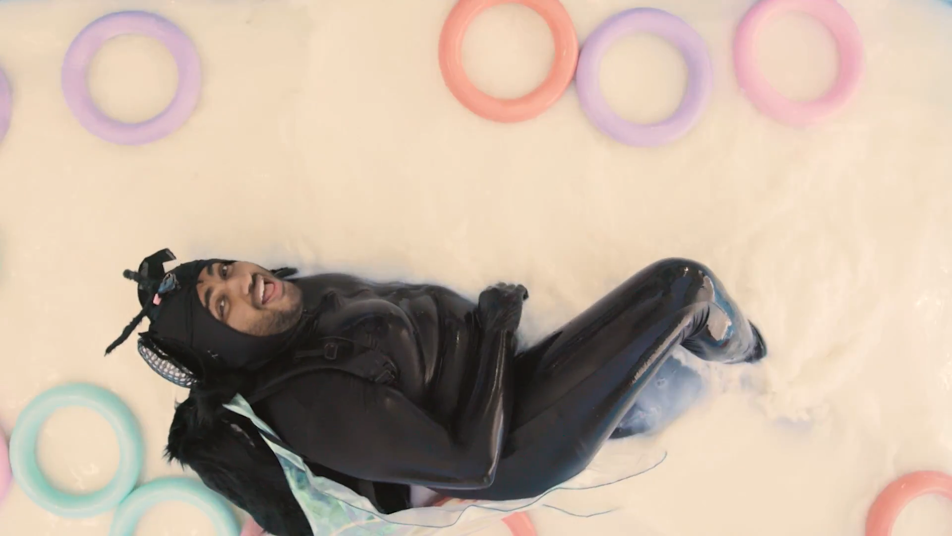 Person in a shiny black outfit lying on a light-colored floor surrounded by pastel inflatable rings