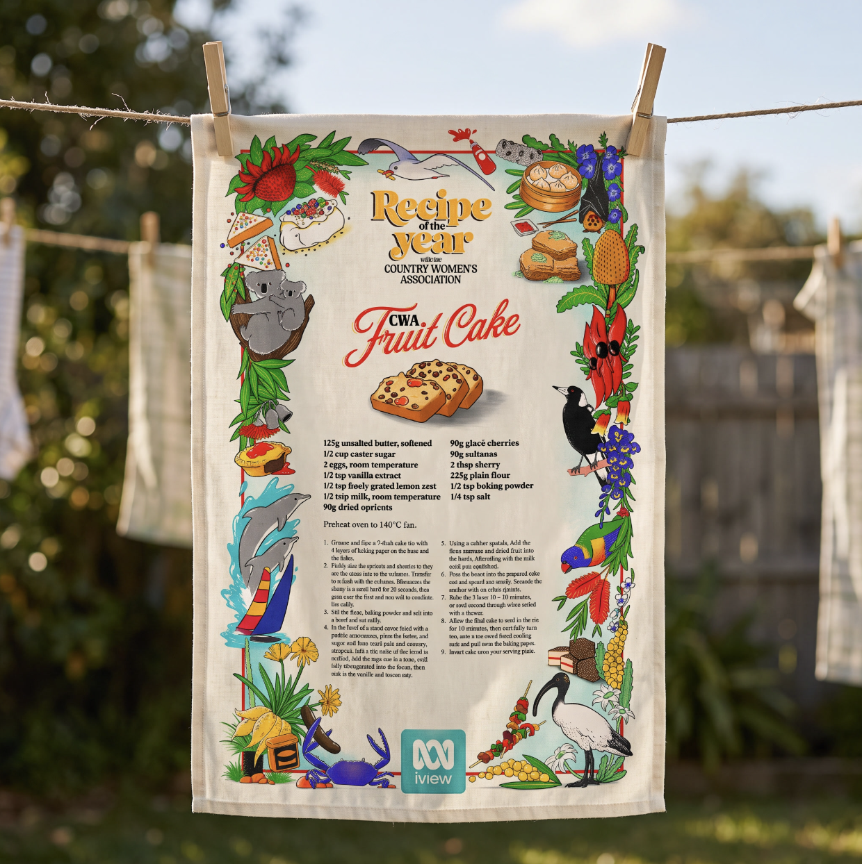 Decorative recipe tea towel hanging on a clothesline, featuring an Australian CWA fruit cake recipe surrounded by colorful native wildlife, flowers, and food illustrations.