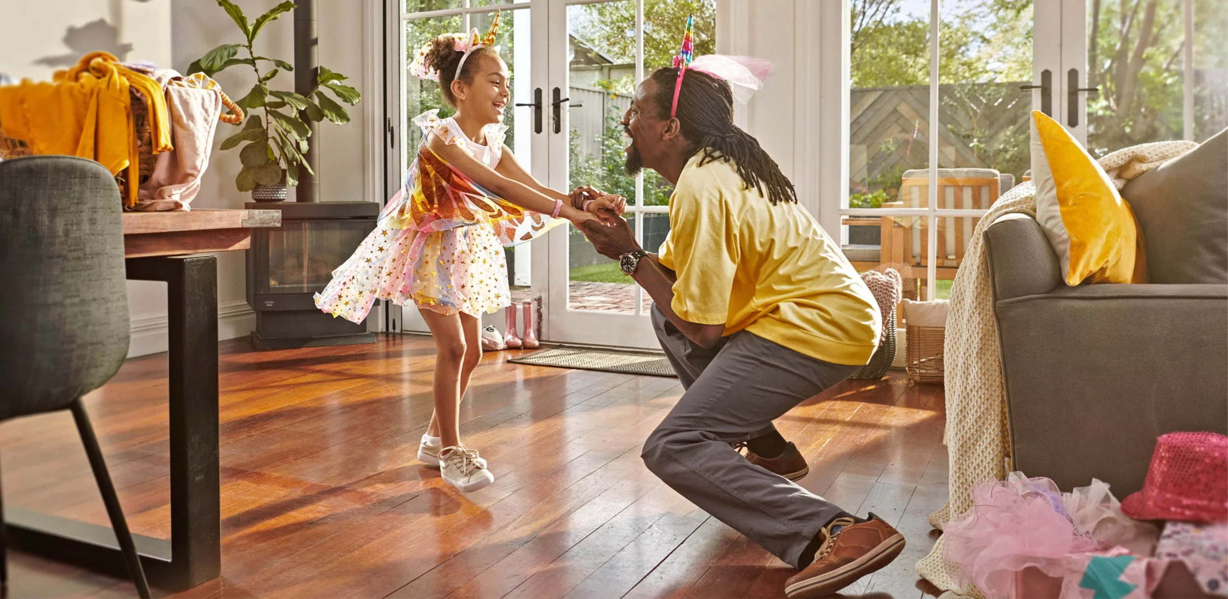 Smiling grandfather kneels and holds hands with a young girl dressed as a fairy in a sunny living room.
