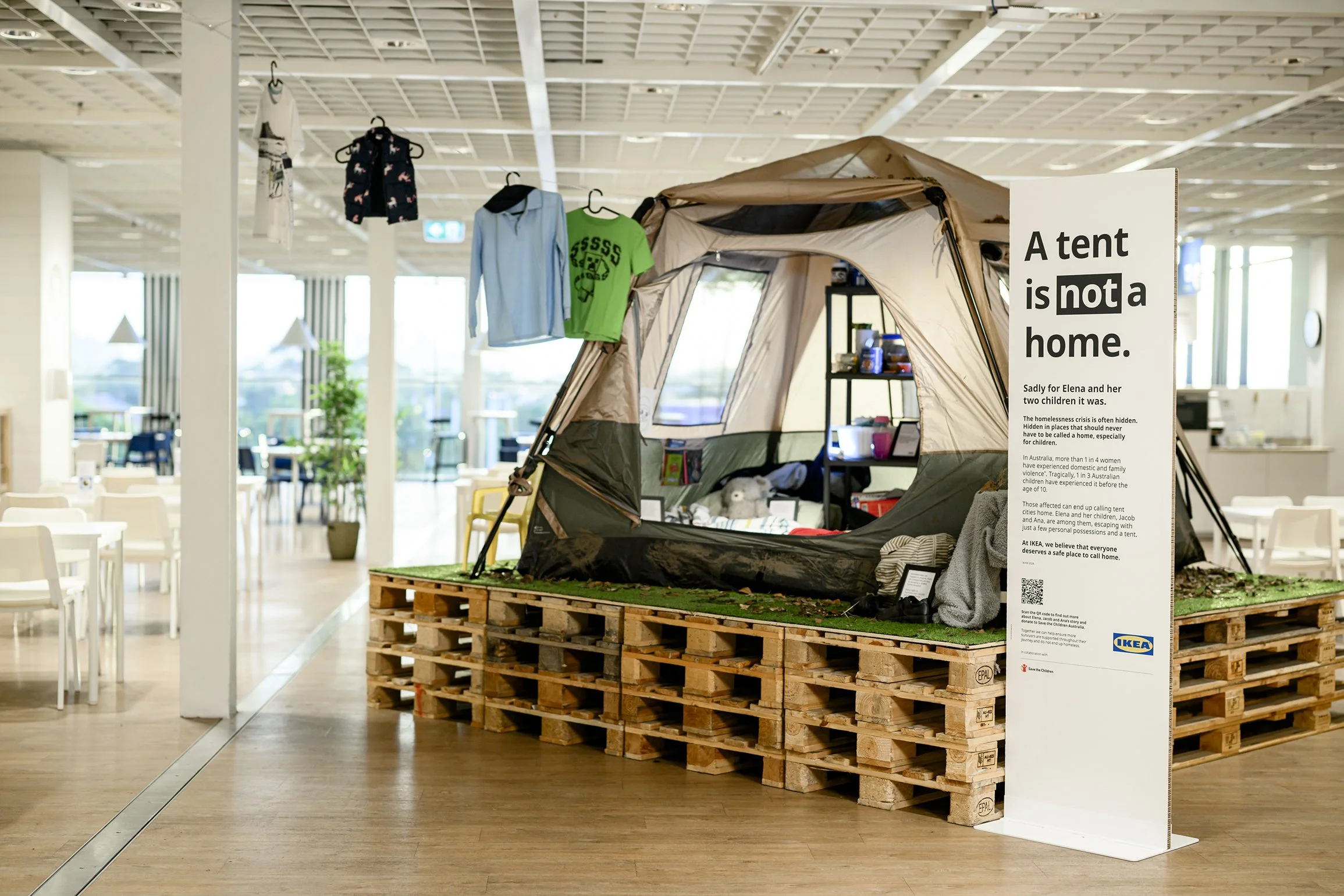 IKEA store display featuring a tent furnished as a family home, hanging clothes, children’s items, and a sign reading “A tent is not a home.”