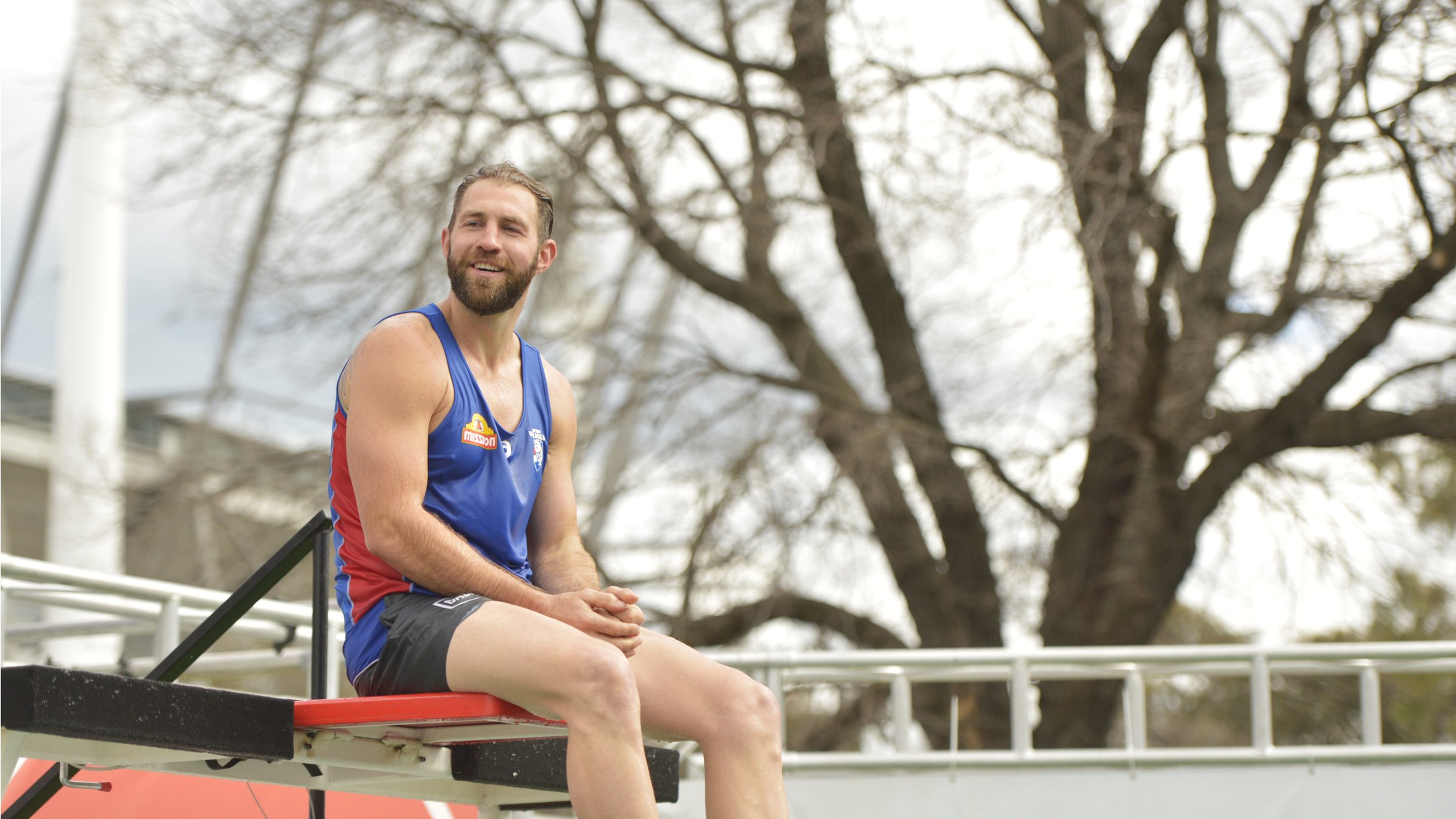 Bearded male athlete in a blue and red rowing uniform sitting on a rowing shell outdoors.