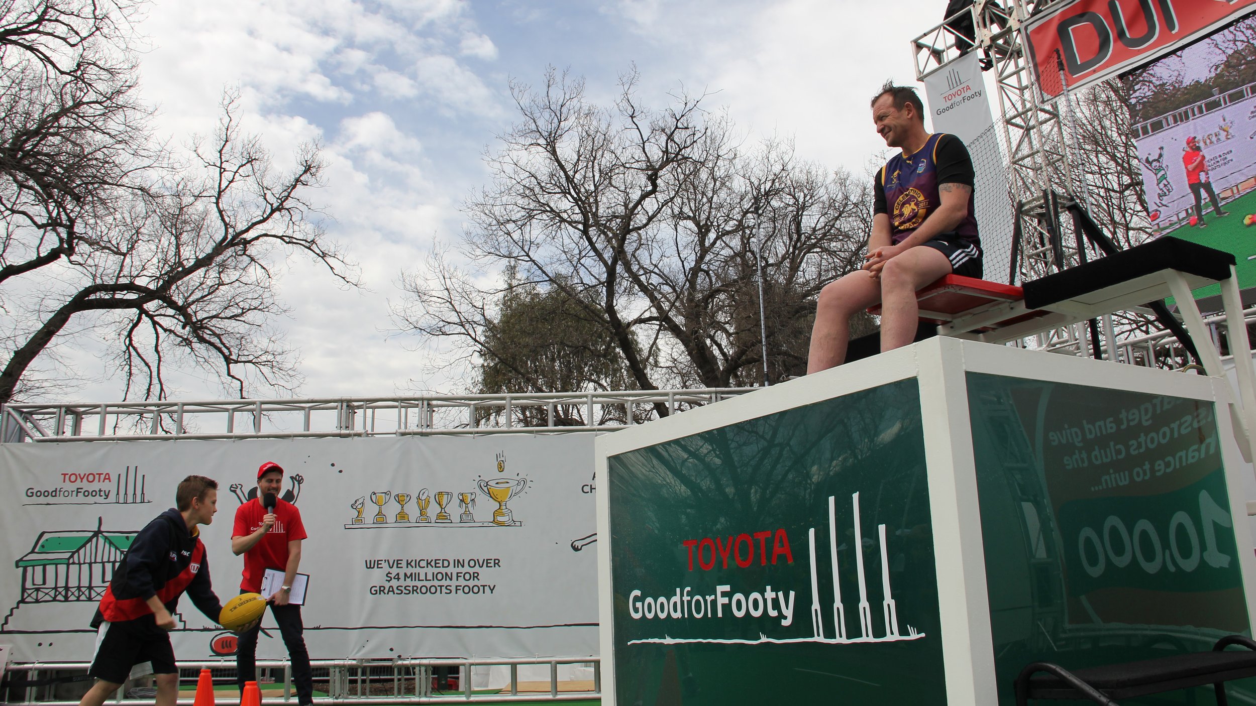Australian rules football promotion with a man seated on a lifeguard-style platform, players handling a football, and Toyota Good for Footy signage.
