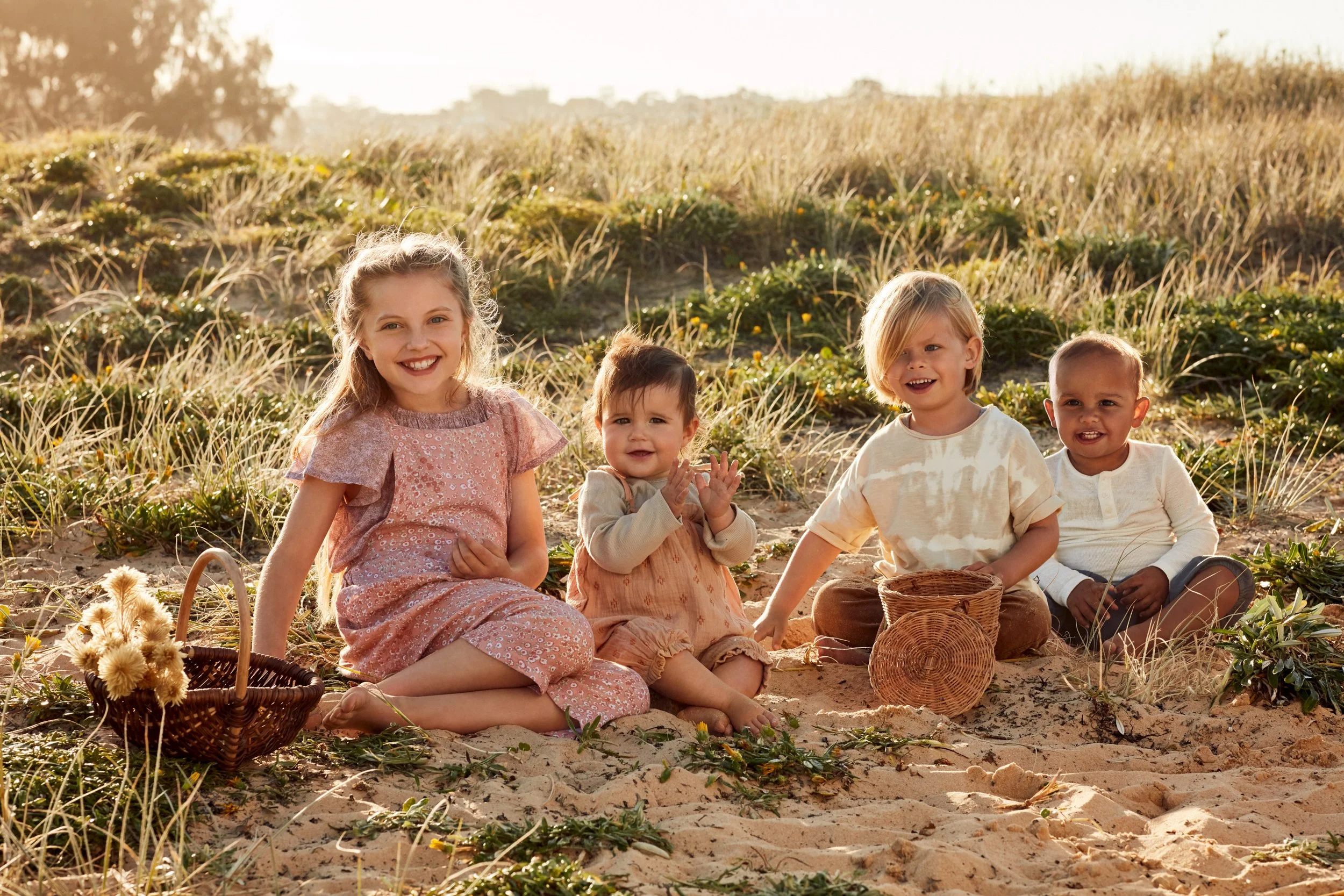 Four children sitting on a sandy beach among dune grass with wicker baskets in warm sunlight