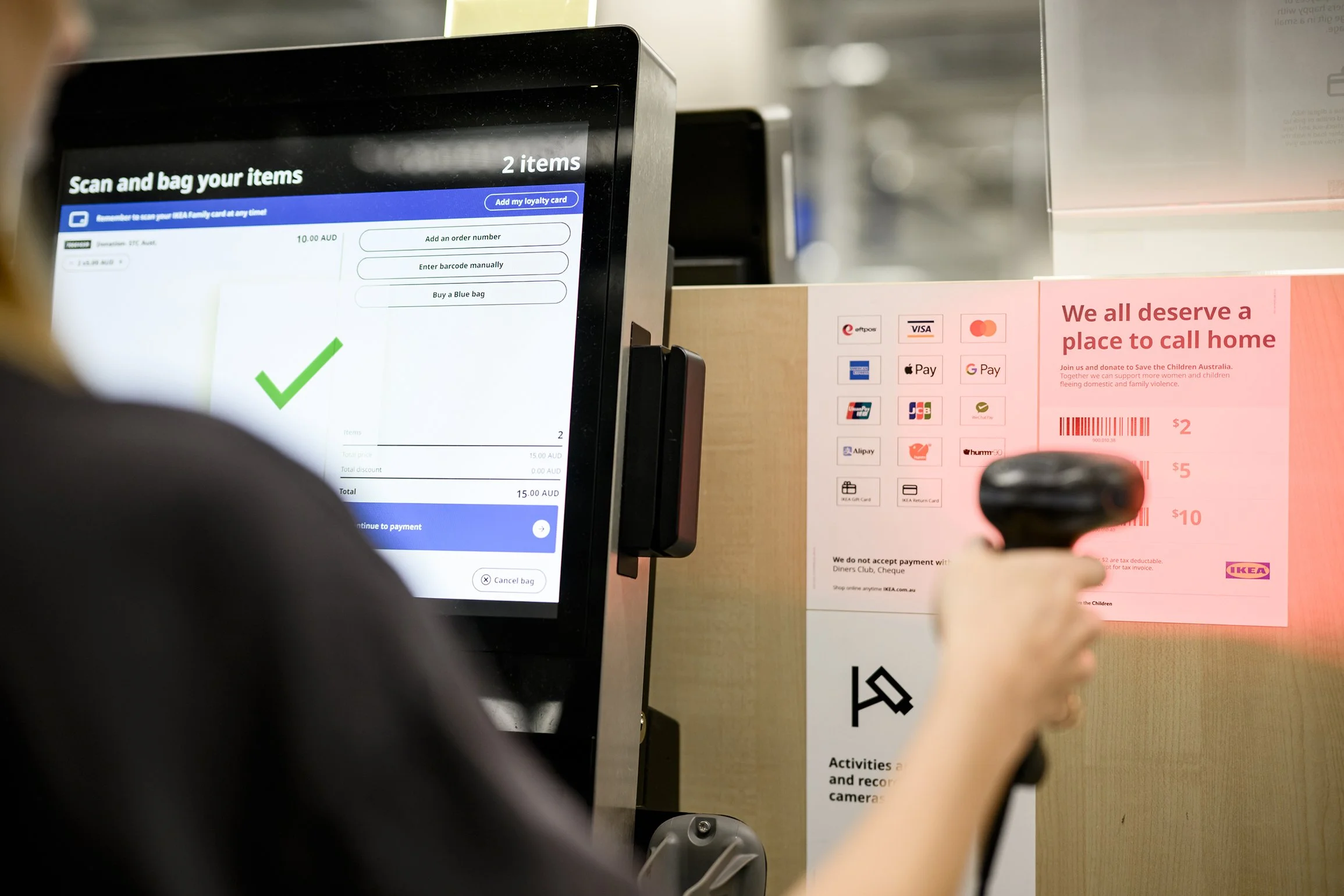 Customer using an IKEA self-checkout kiosk beside a payment options and charity donation sign.