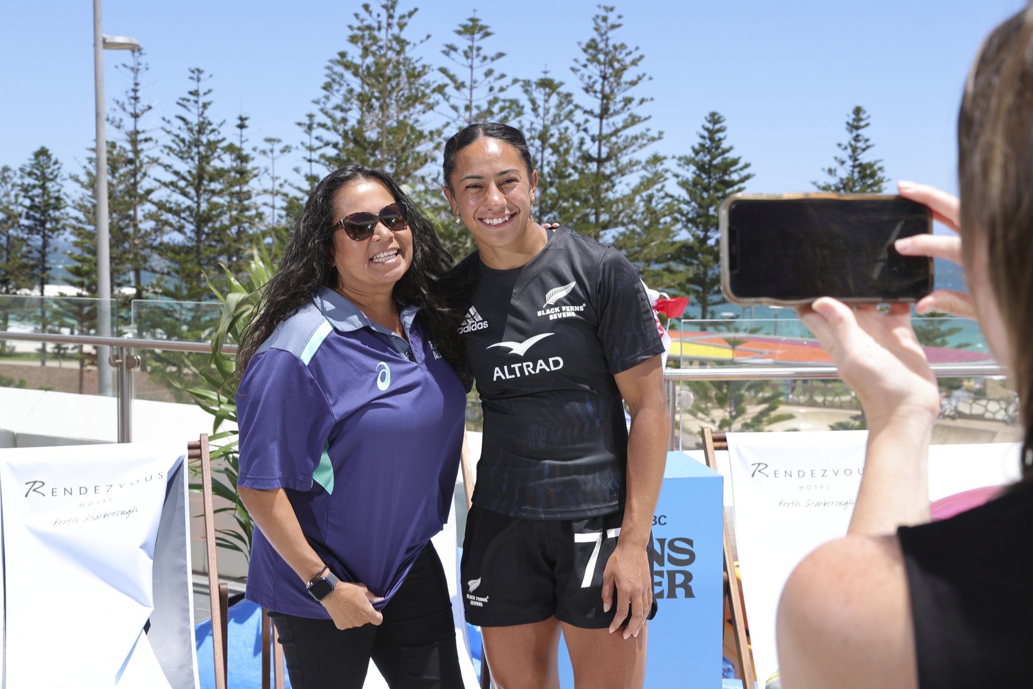 Two smiling women pose outdoors while a person photographs them with a smartphone.