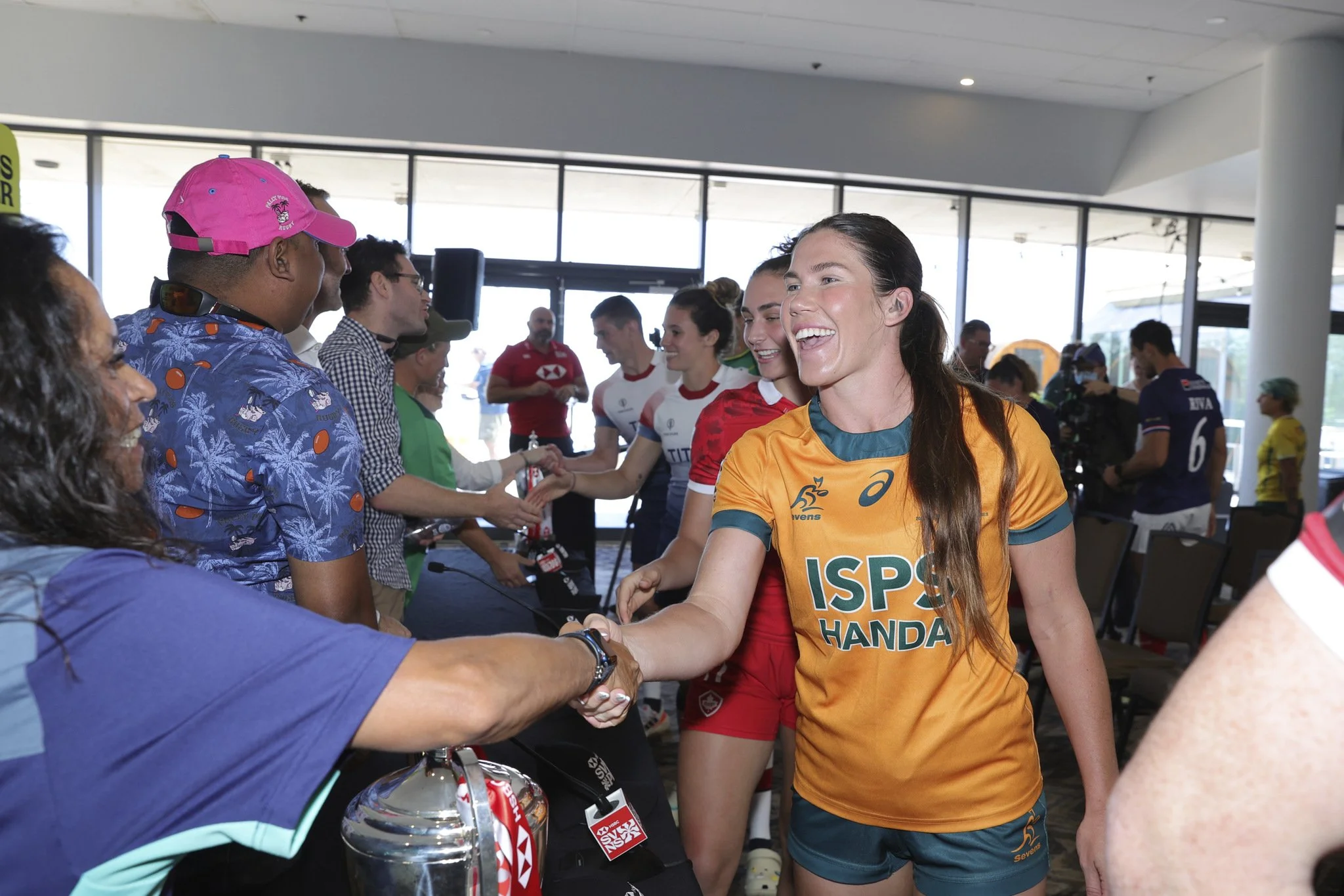 Smiling Australian rugby player in an orange ISPS Handa jersey shakes a fan’s hand at an indoor meet-and-greet.
