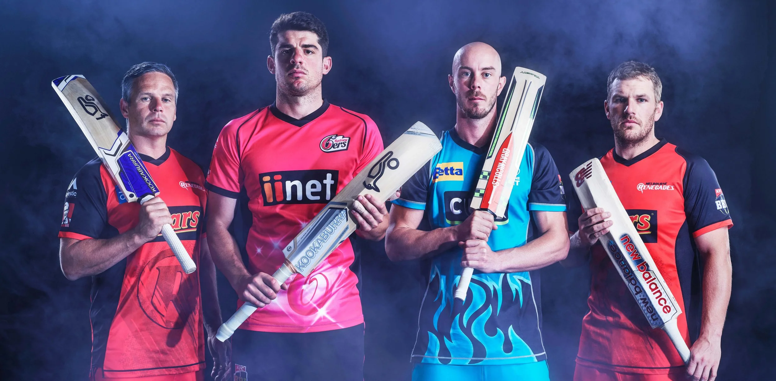 Four professional cricket players posing with bats in colorful team uniforms against a smoky blue background