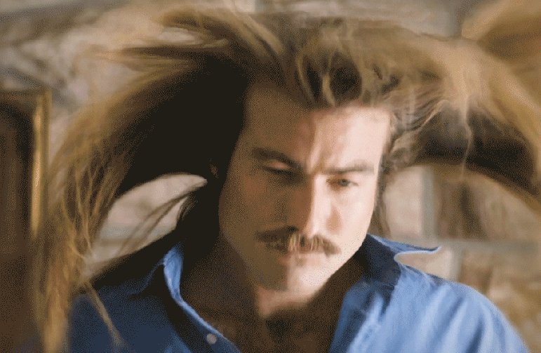 Close-up portrait of a mustached man in a blue shirt with windblown hair outdoors