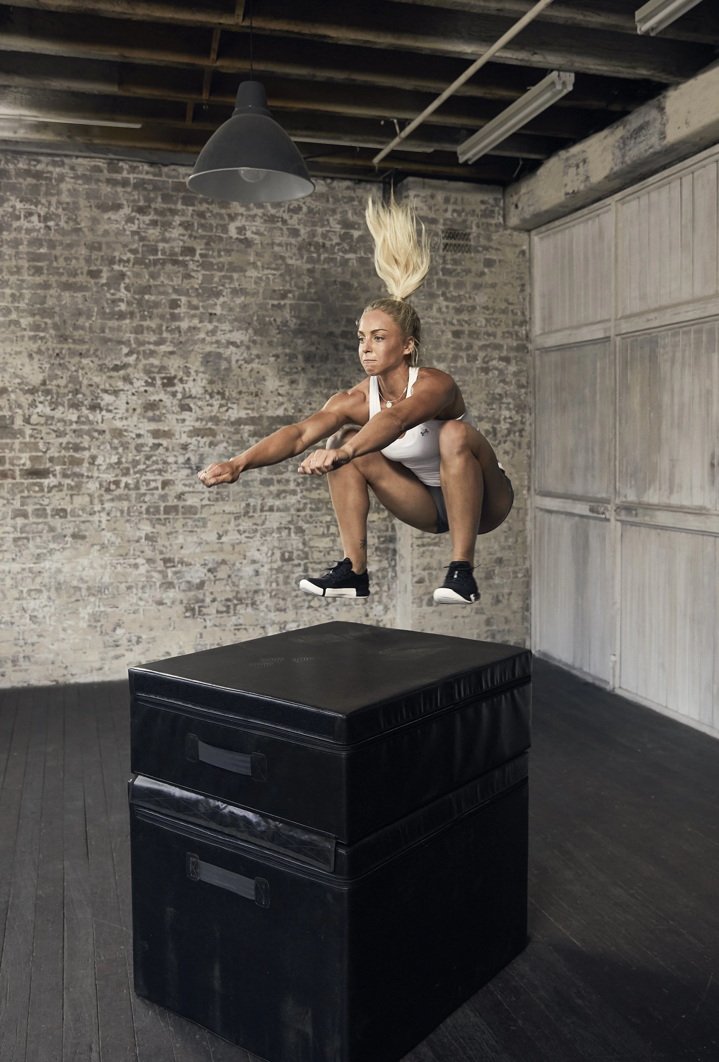 Woman performing a box jump onto a black plyometric platform in an industrial-style gym.